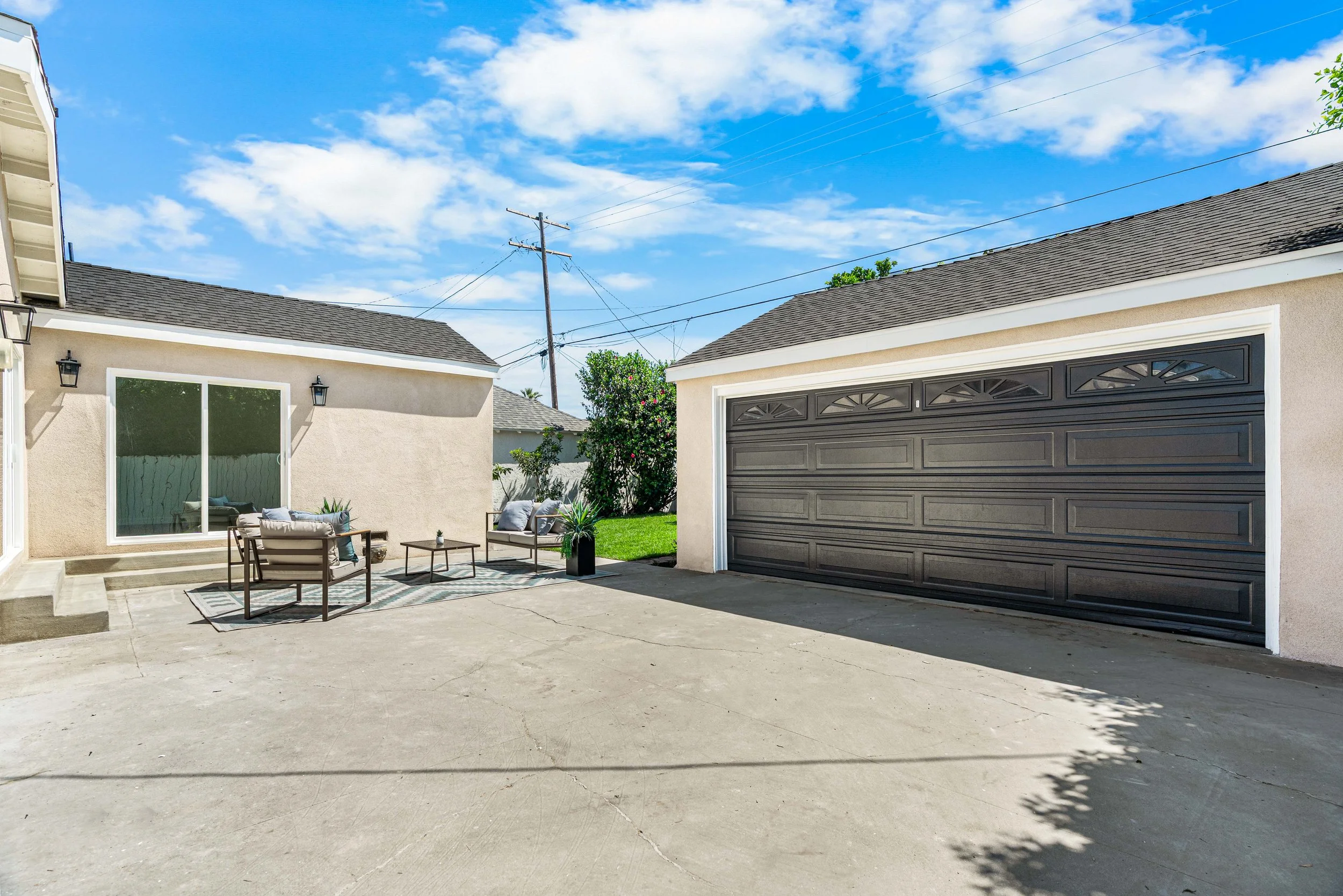 A backyard patio with outdoor furniture, including chairs, a small table, a rug, and decorative pillows, surrounded by a beige house with large windows and a gray garage door. There are trees, a grassy area, a wooden fence, and a blue sky with scatte
