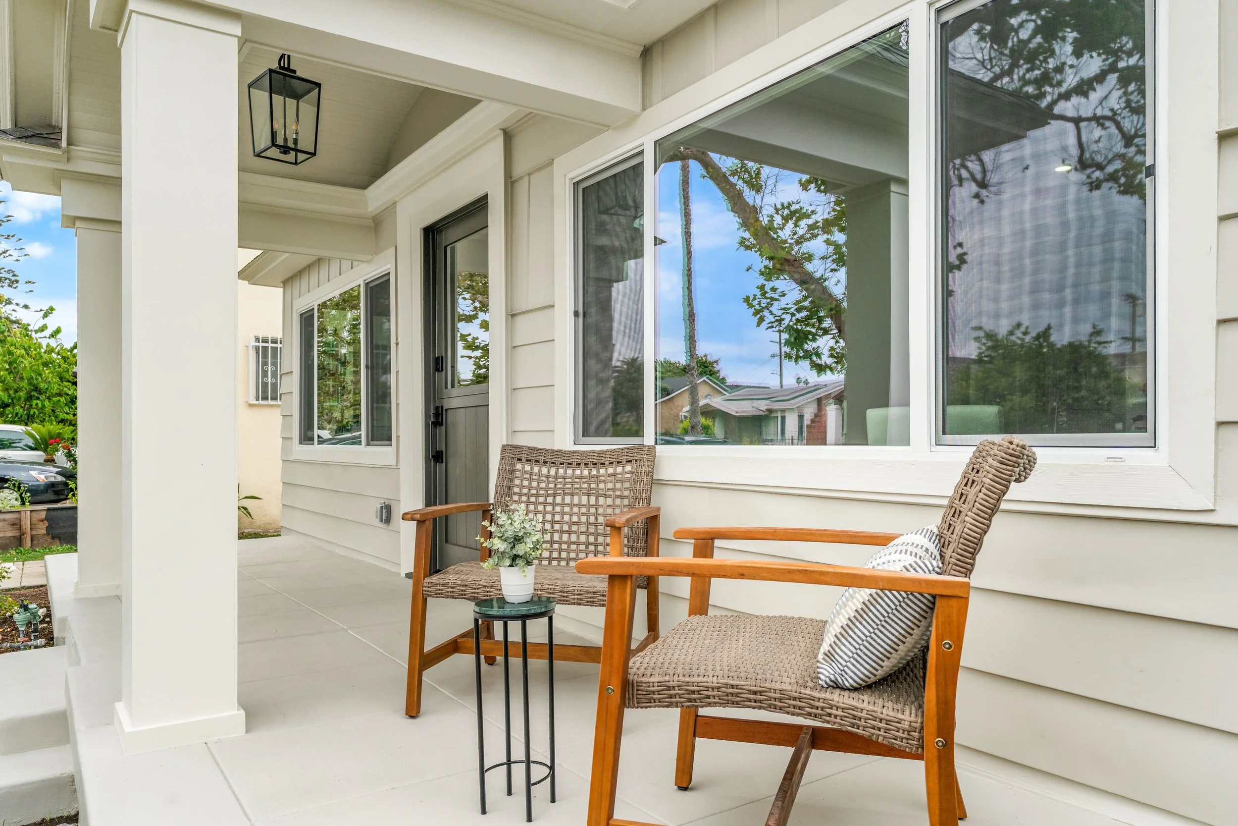 A front porch with two wooden chairs, a small side table with a flower pot, and large windows reflecting trees and sky. There is a hanging lantern on the ceiling and a gray door on the wall.