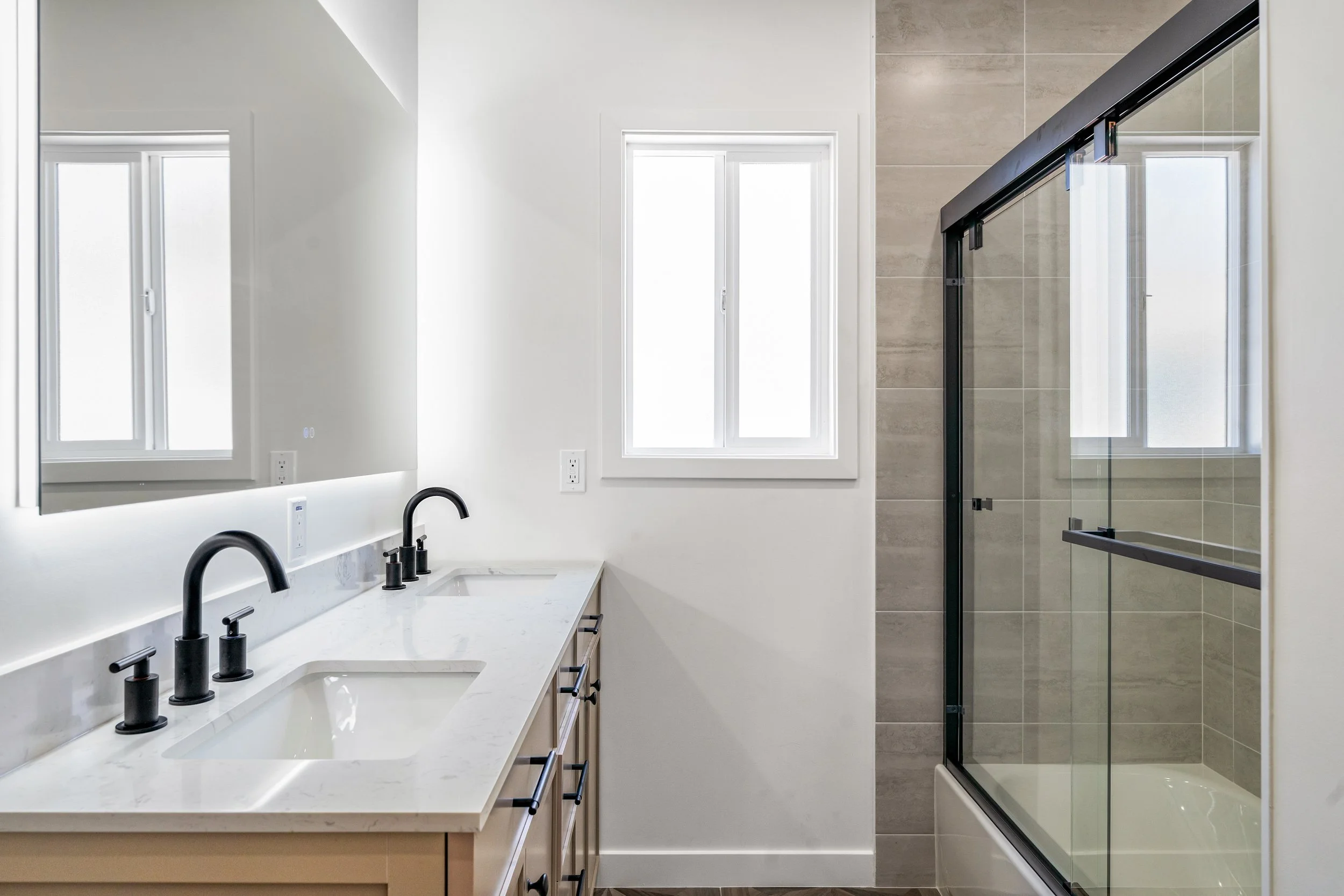 Modern bathroom with a white marble countertop, double sinks with black faucets, and a large mirror. There are two windows providing natural light, and a glass shower enclosure with beige tiled walls.