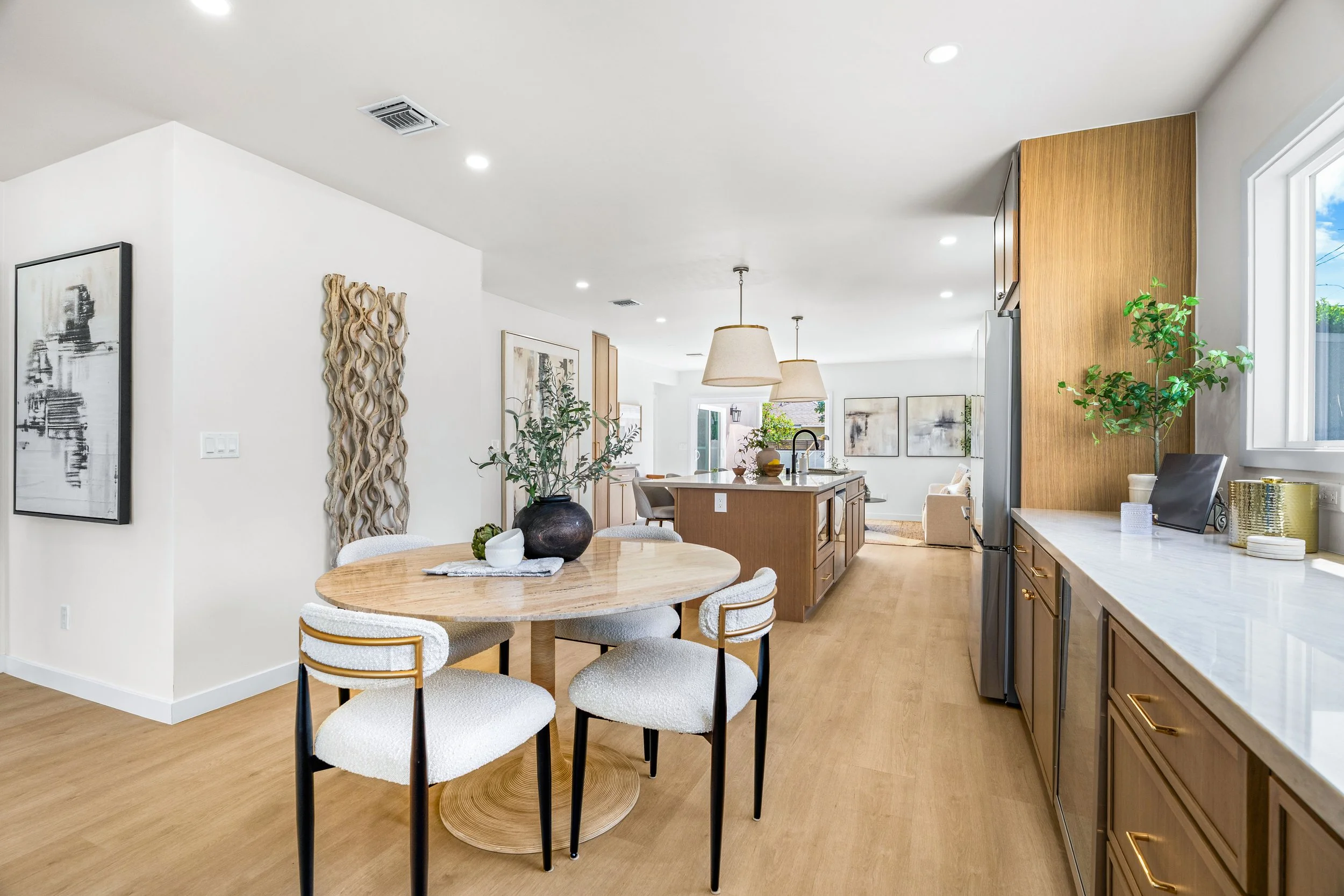 Bright, modern kitchen and dining area with a round wooden table and white chairs. Kitchen features wooden cabinets, a marble countertop, and green plants near a window.