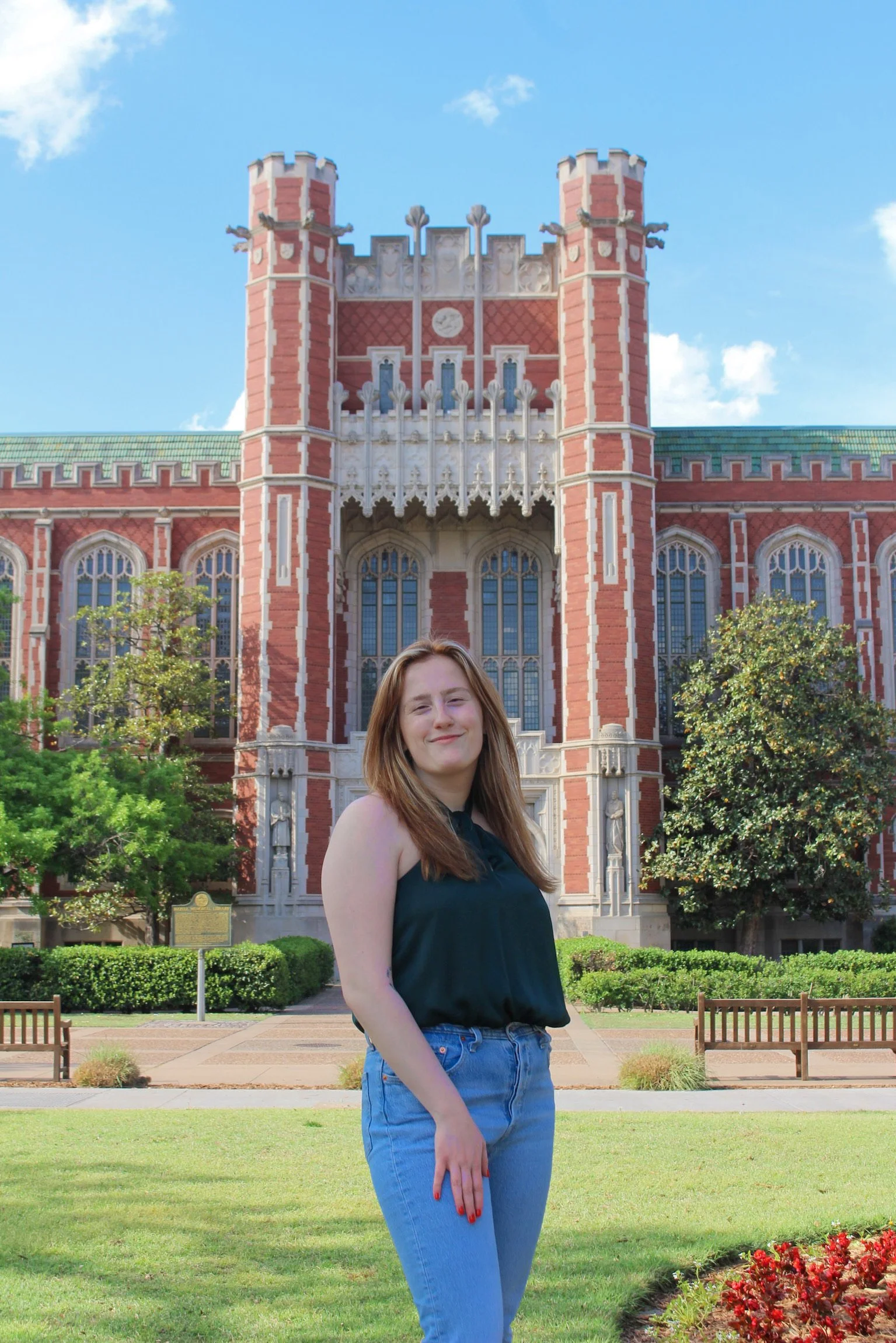 A woman with long reddish-brown hair smiling, standing outside in front of a historic red brick building with ornate windows and towers, greenery, a flower bed, until a blue sky with a few clouds.