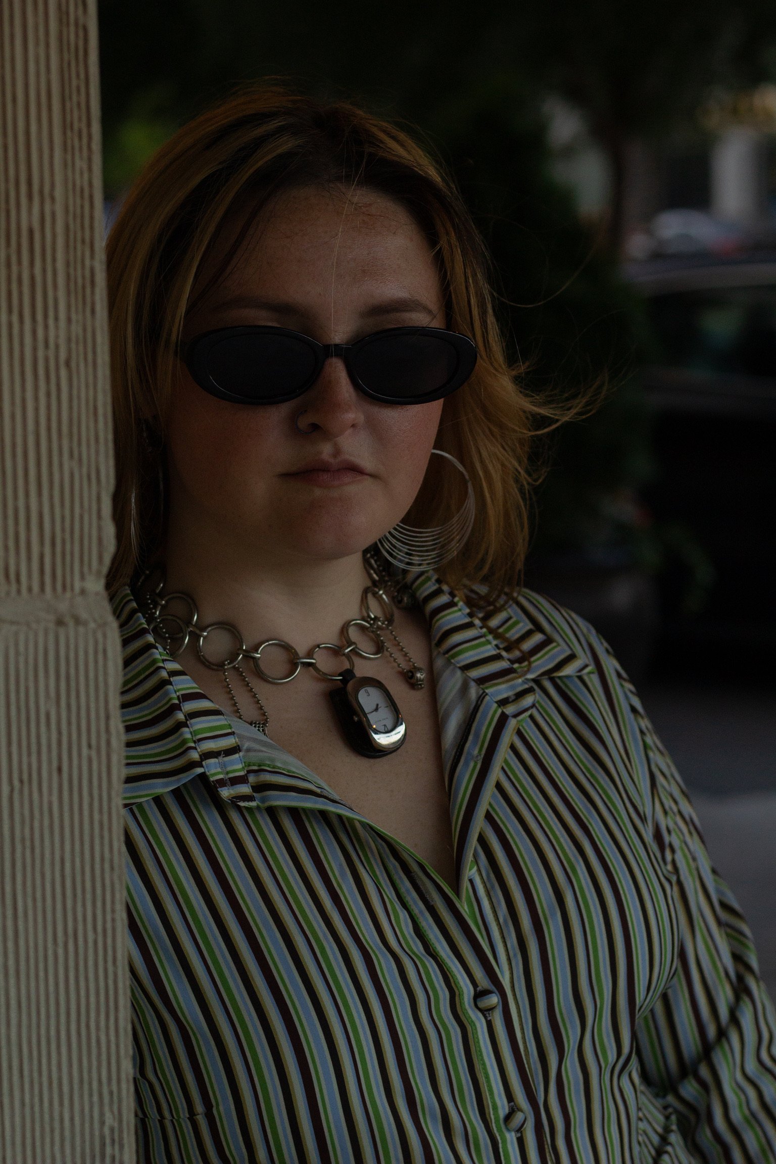 A woman with light skin and wavy, shoulder-length blonde hair, wearing dark sunglasses, a striped shirt, and layered necklaces, standing outdoors near a wooden post.