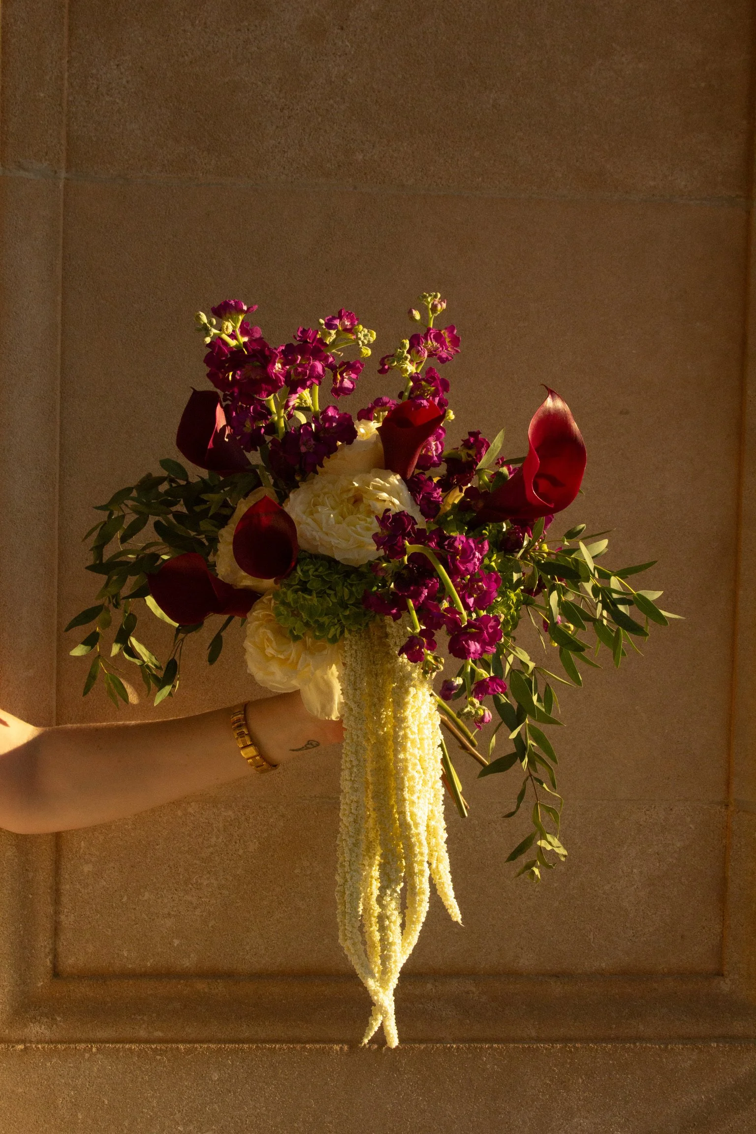 A person wearing a gold bracelet and a ring, holding a floral arrangement with red, white, purple, and green flowers, including roses and calla lilies, on a beige textured background.