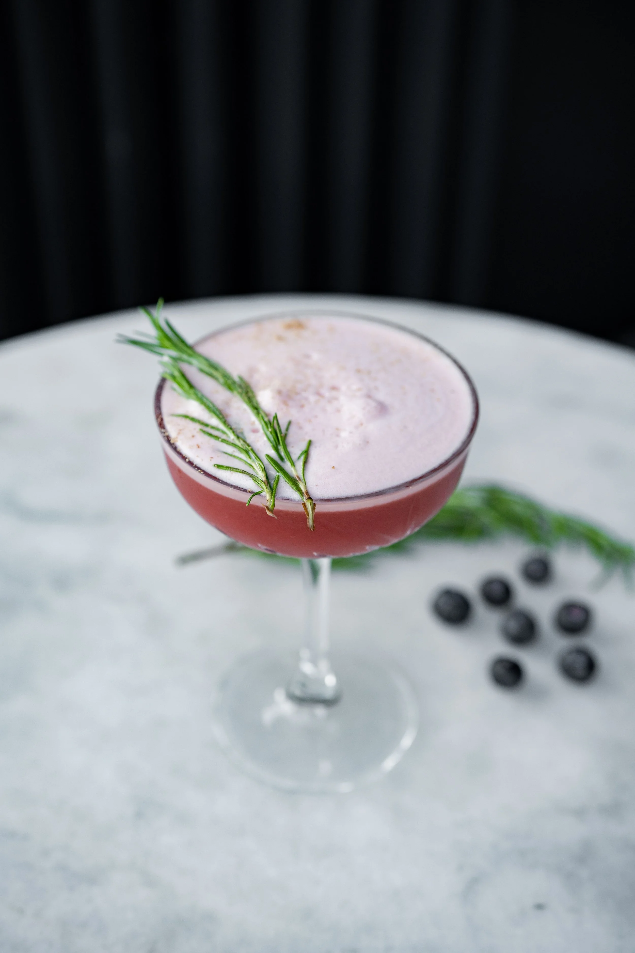 A pink cocktail with a frothy top and a sprig of rosemary in a coupe glass, with blackberries scattered on a white surface around it and a dark background.