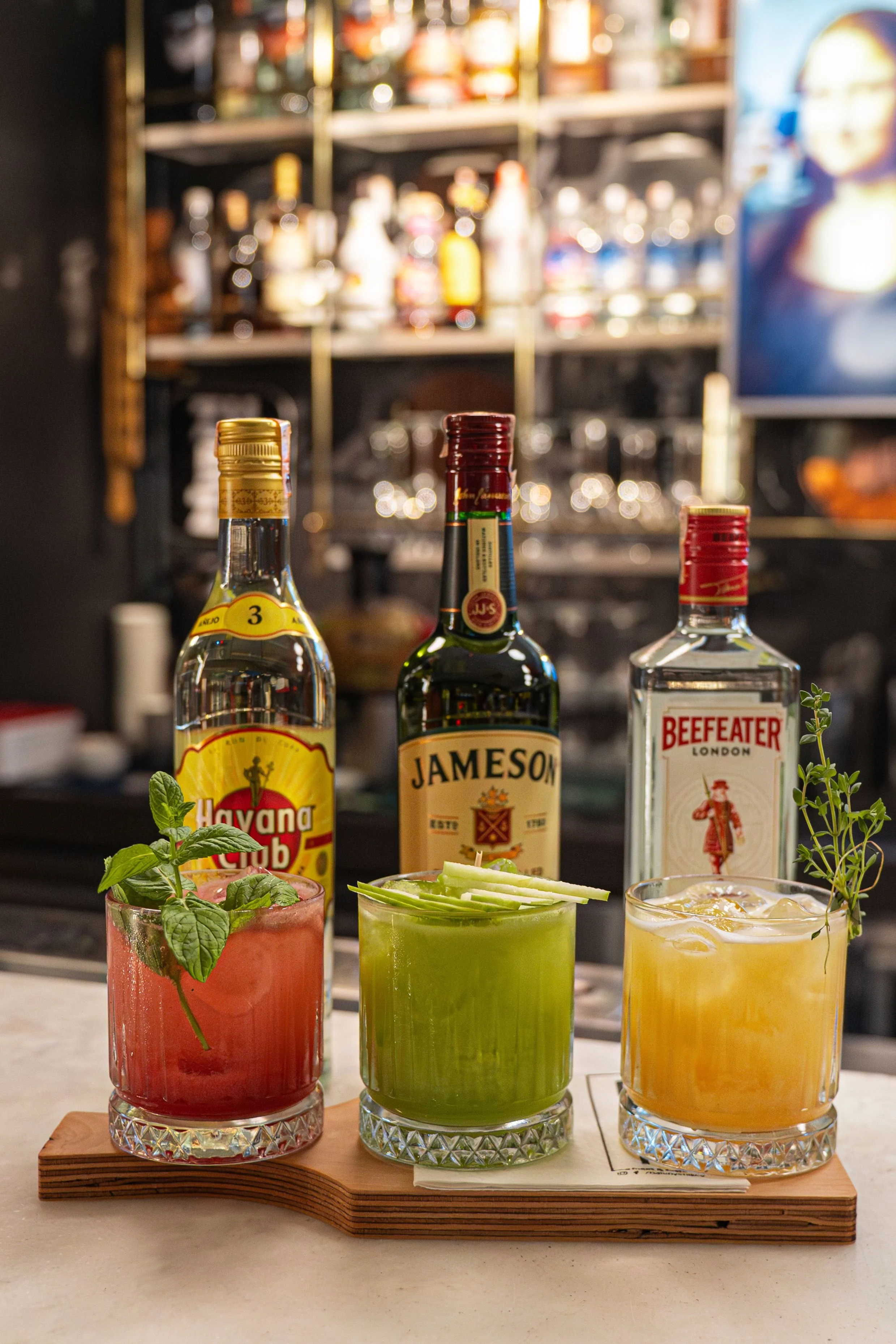 Three colorful cocktails garnished with fresh herbs on a wooden serving board, with liquor bottles blurred in the background.