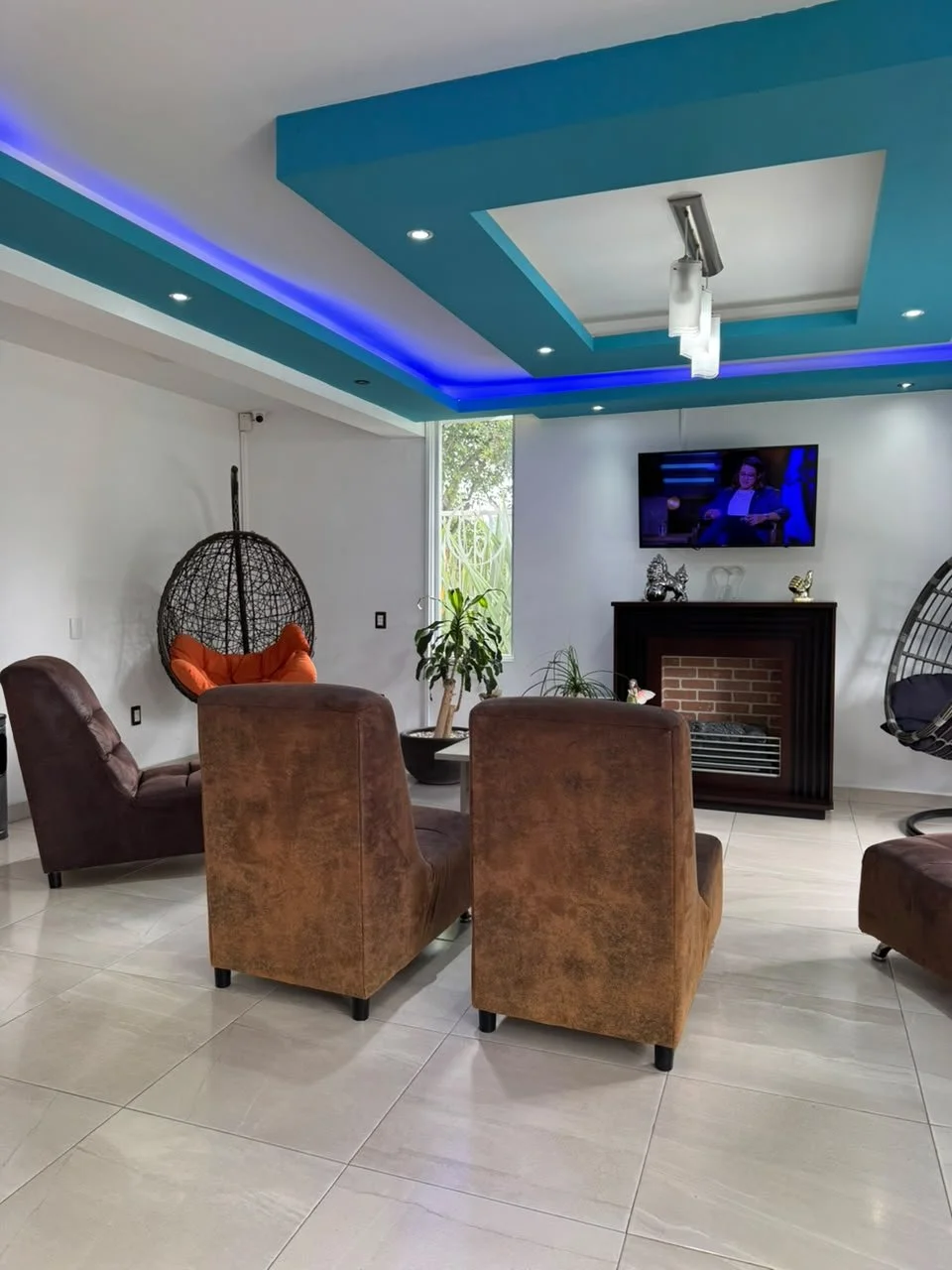 Sala moderna con sillones de tela marrón, una silla columpio negra, plantas, una chimenea eléctrica y una televisión en la pared con luces azules en el techo.