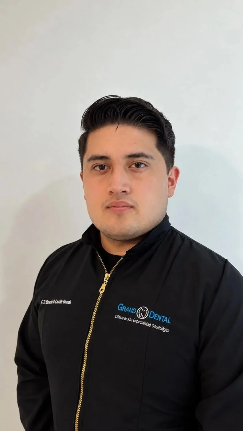 Joven con cabello oscuro y apariencia seria, usando chaqueta negra con logotipo de clínica dental y texto en español, posando frente a una pared blanca.