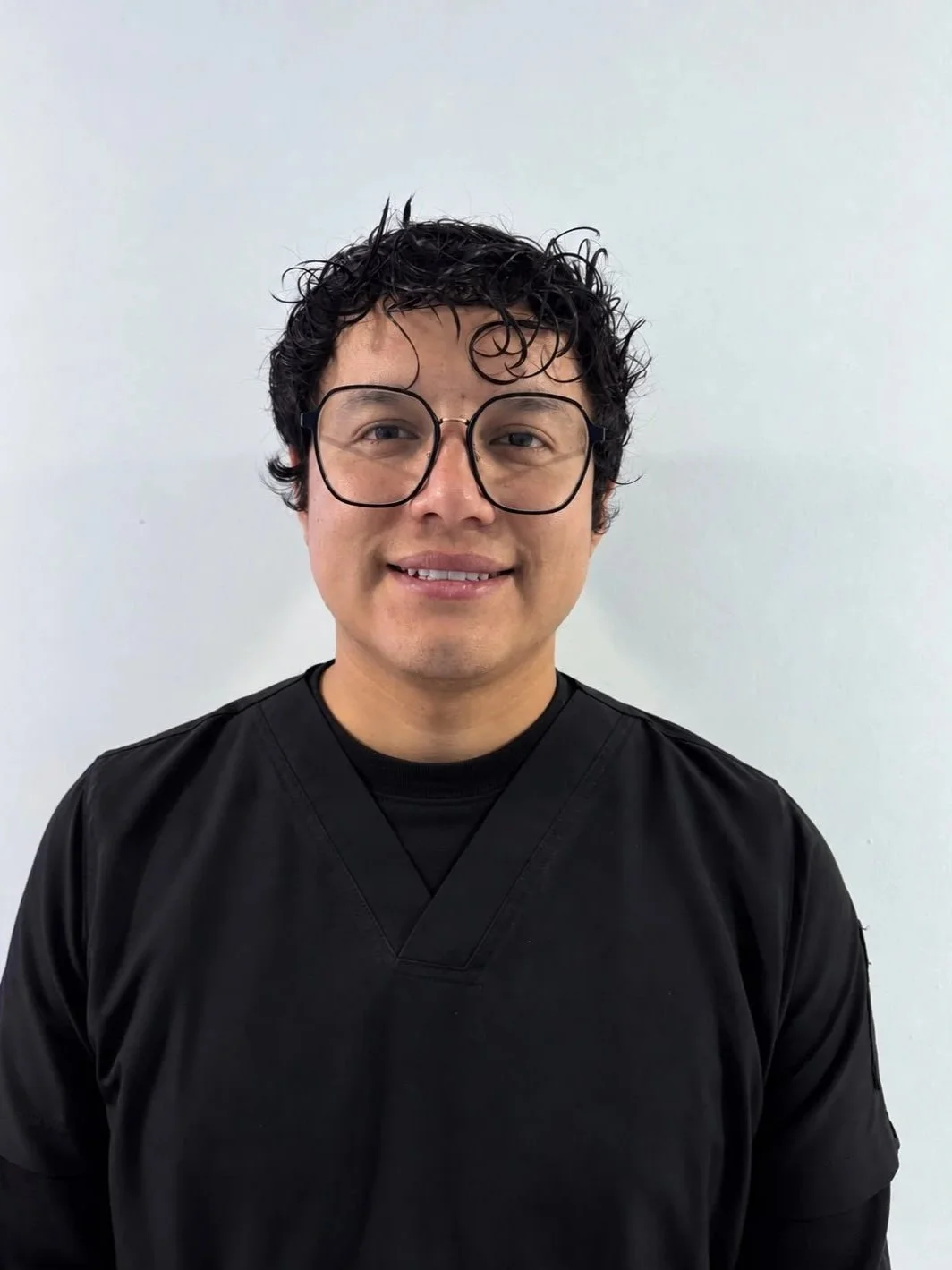 Hombre con gafas y cabello rizado usando ropa negra frente a un fondo blanco.