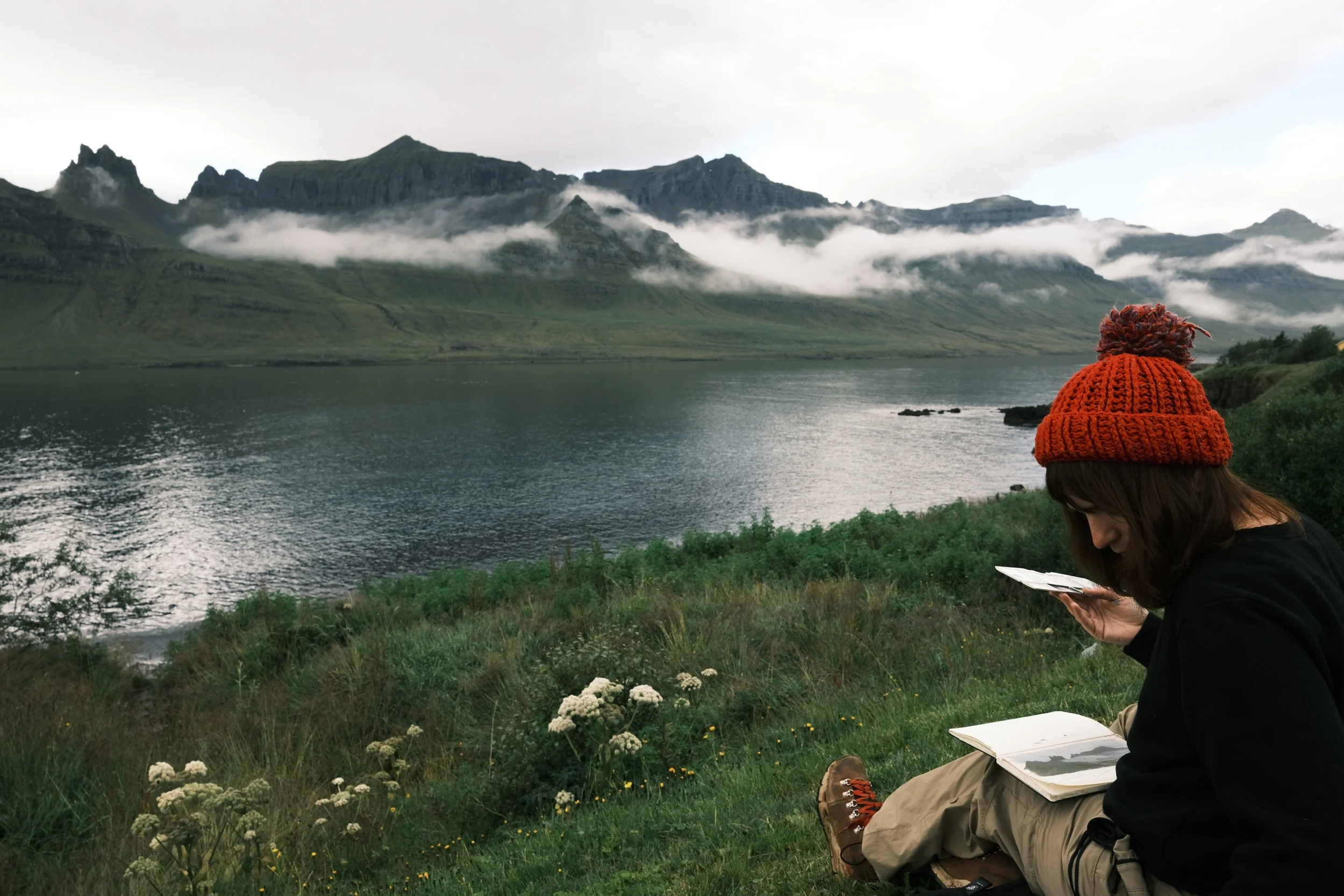 A person with a red knit hat sitting on grass near a lake, reading a book, with mountains and clouds in the background.