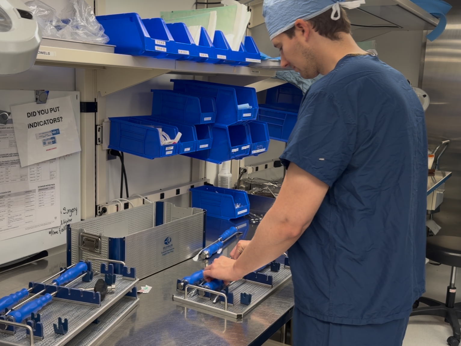 A healthcare worker in blue scrubs and cap working at a hospital supply station with blue bins and surgical tools on a workbench.
