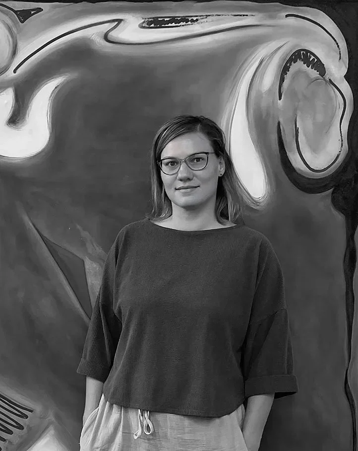 A woman standing in front of a large abstract graffiti mural, wearing glasses, a loose dark shirt, and light-colored pants.