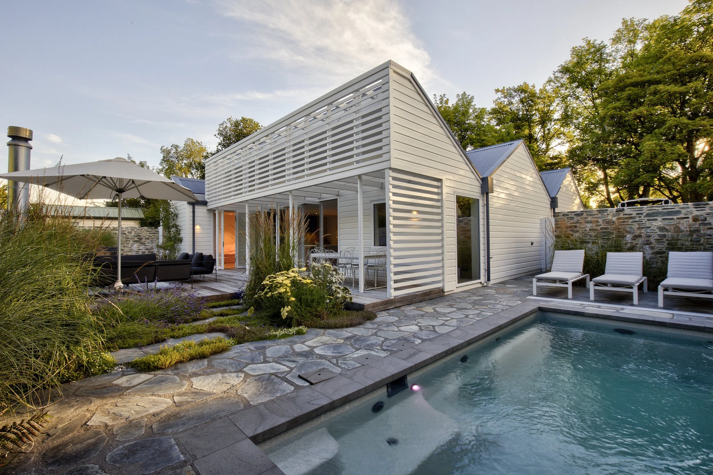 A modern white house with a swimming pool in the backyard, outdoor lounge chairs, an umbrella, and surrounding greenery during the evening.