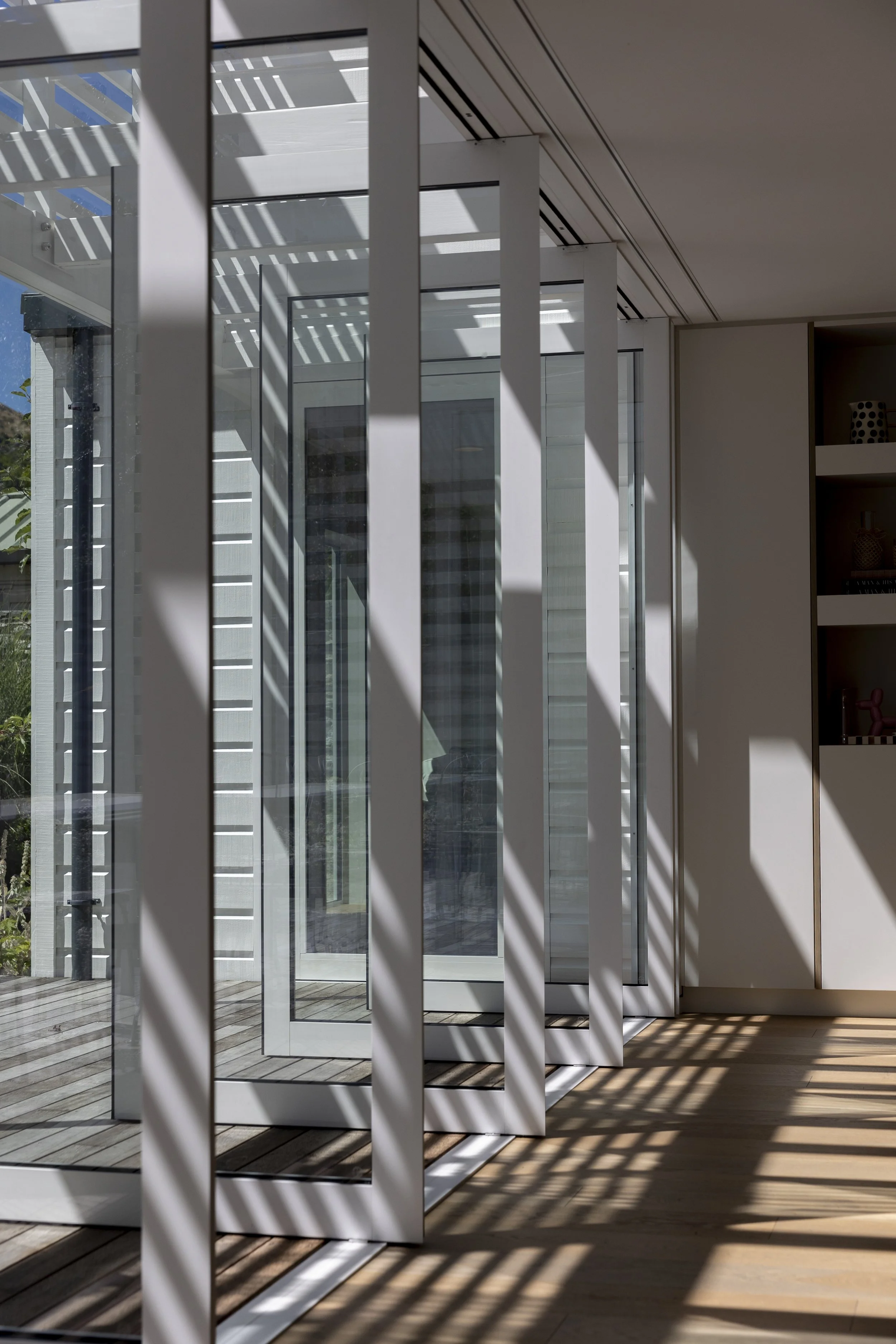 Indoor scene showing a glass door, with sunlight creating shadow patterns on a wooden floor. The door is partially open, revealing an outdoor balcony with a pergola.