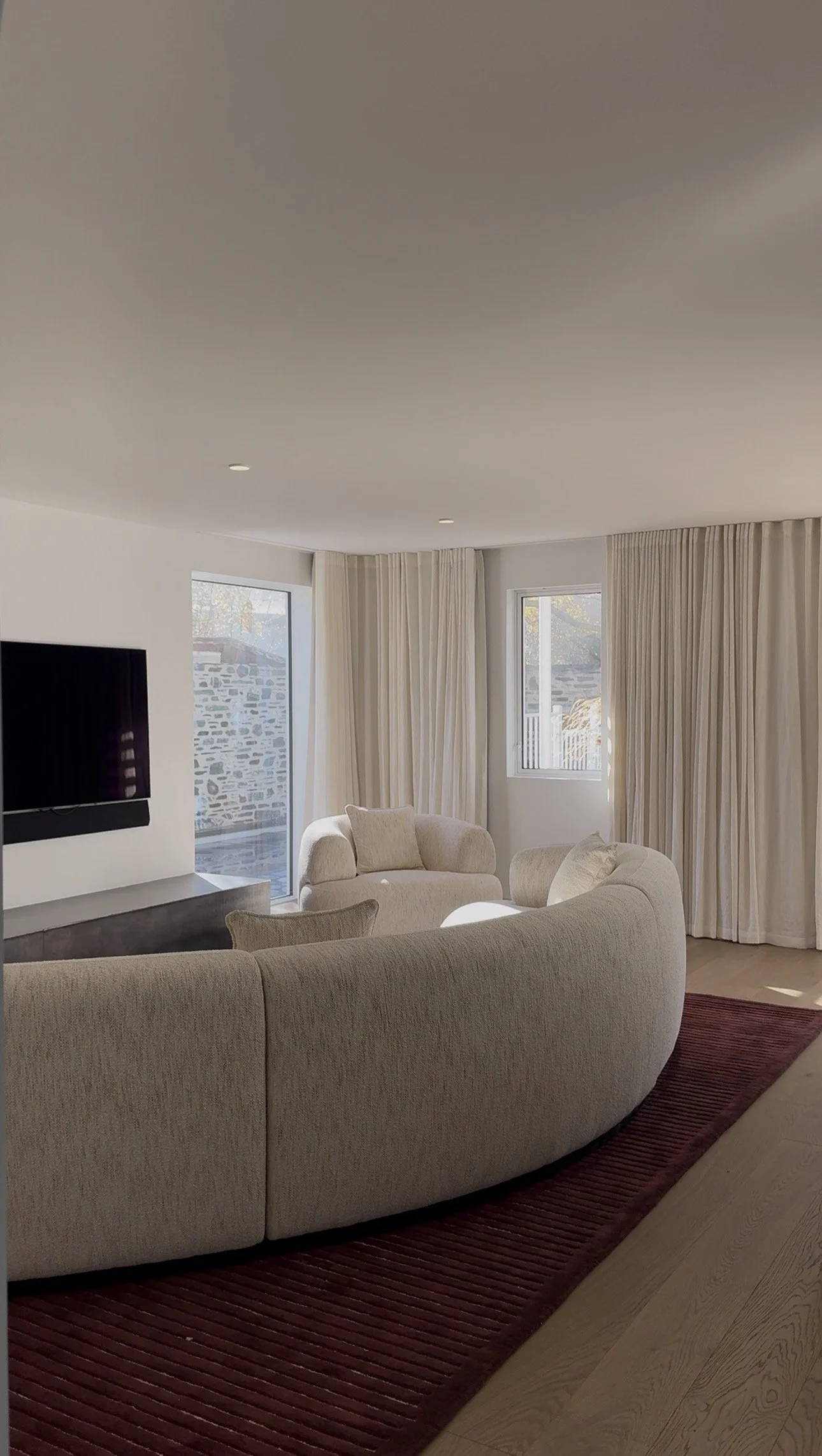 Living room with a curved beige sofa, white armchairs, a wall-mounted flat-screen TV, with windows and beige curtains, a dark red rug, and wooden flooring.