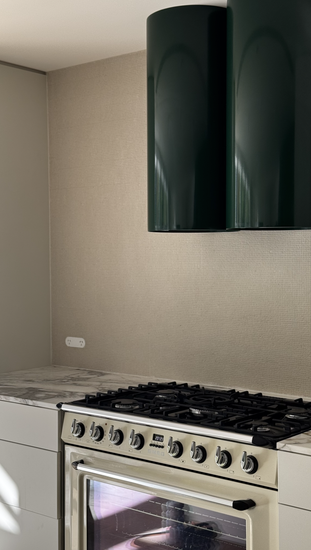 A modern kitchen with a beige textured wall, a white oven with metallic knobs, a marble countertop, and dark green wall-mounted cabinets.