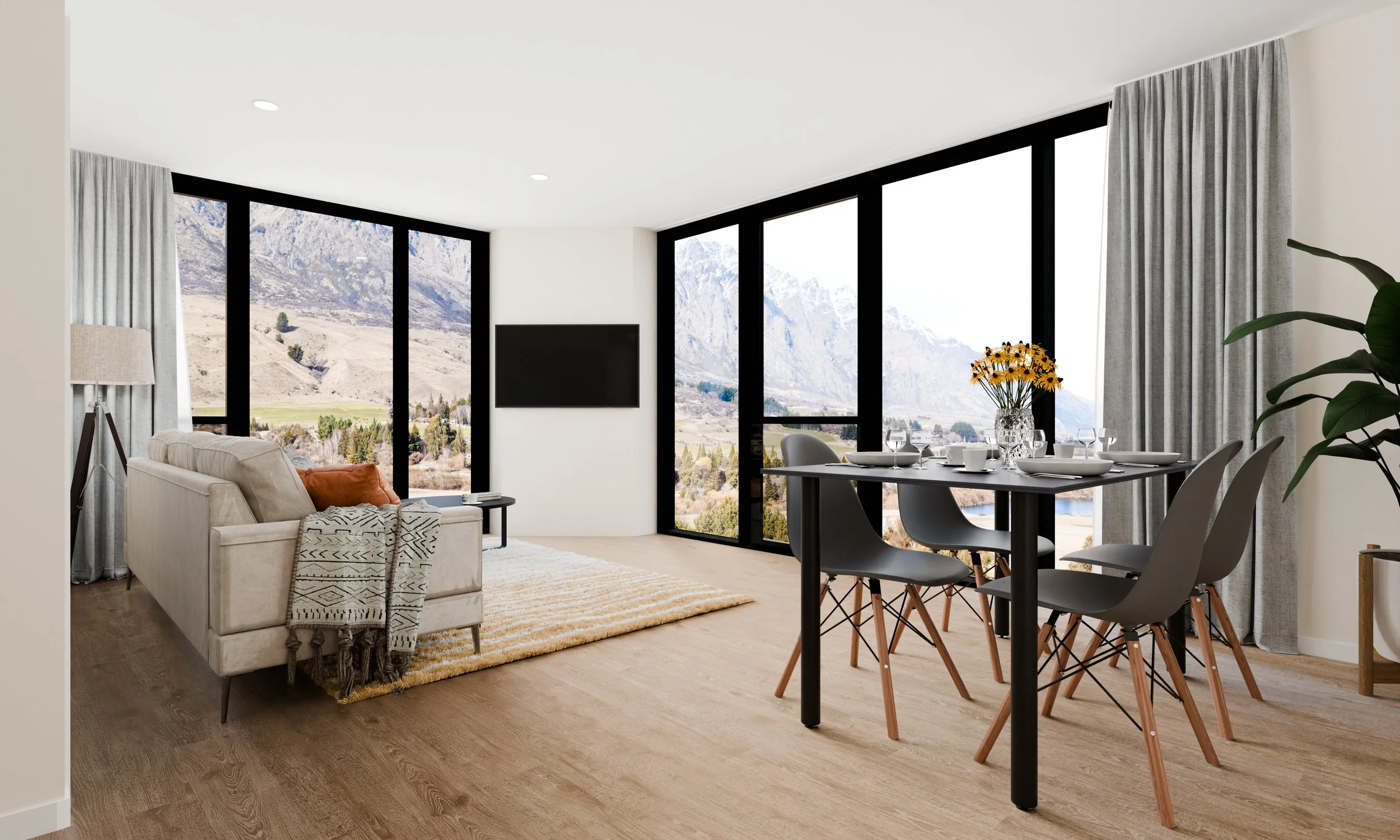 Living and dining area with large floor-to-ceiling windows showing mountainous landscape, a beige sofa with throw pillows, a dining table set with white dishes and glassware, a vase with yellow flowers, and a tall green plant.