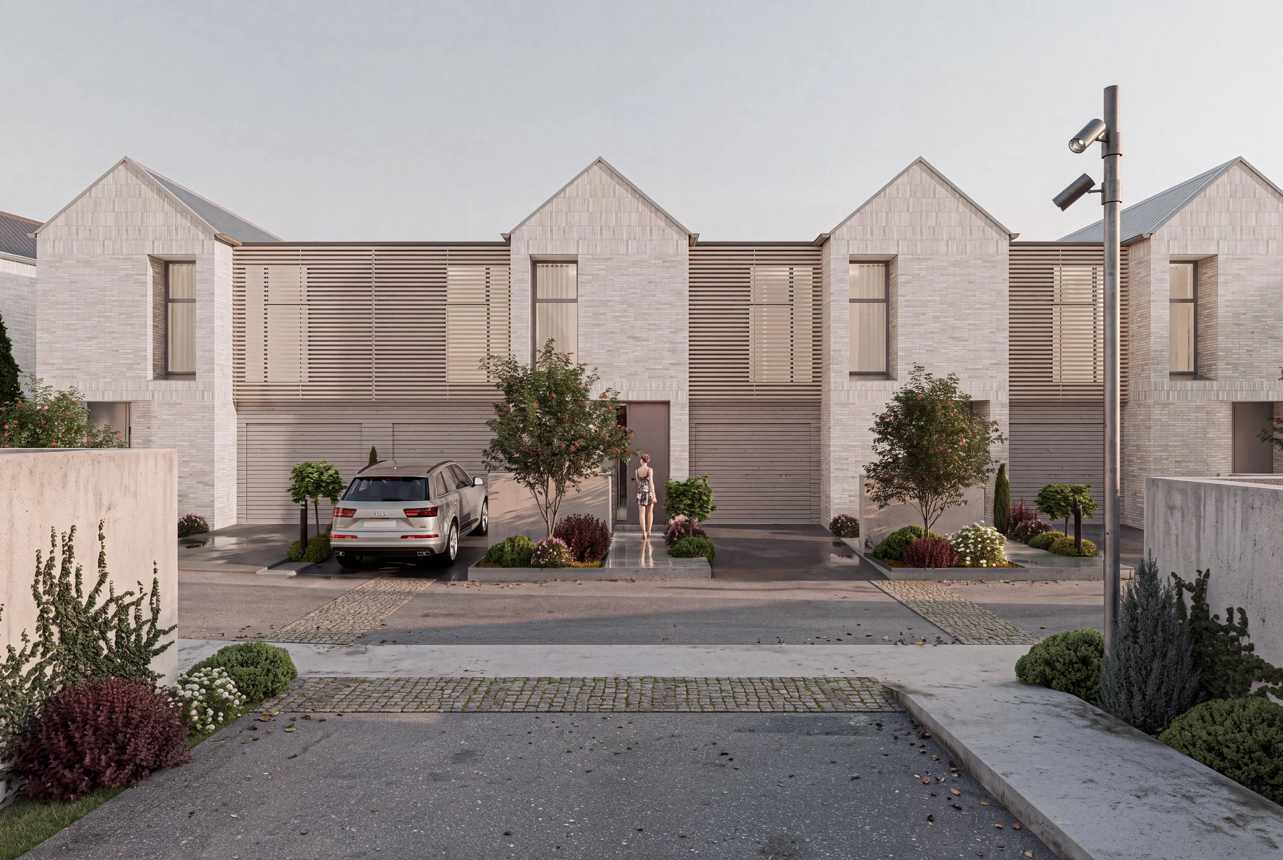 Modern row of townhouses with light-colored brick exteriors and gabled roofs, landscaped front yards with bushes and trees, parked car, person walking, and streetlamp in foreground.