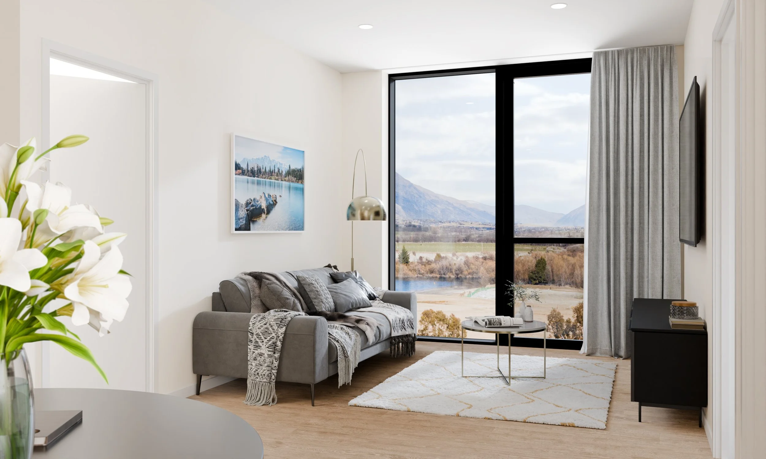 Living room with a large window offering mountain and lake views, a gray sofa with pillows and a throw blanket, a white area rug, a small round coffee table, a wall-mounted TV, a modern floor lamp, a framed landscape picture, and a vase of white flow