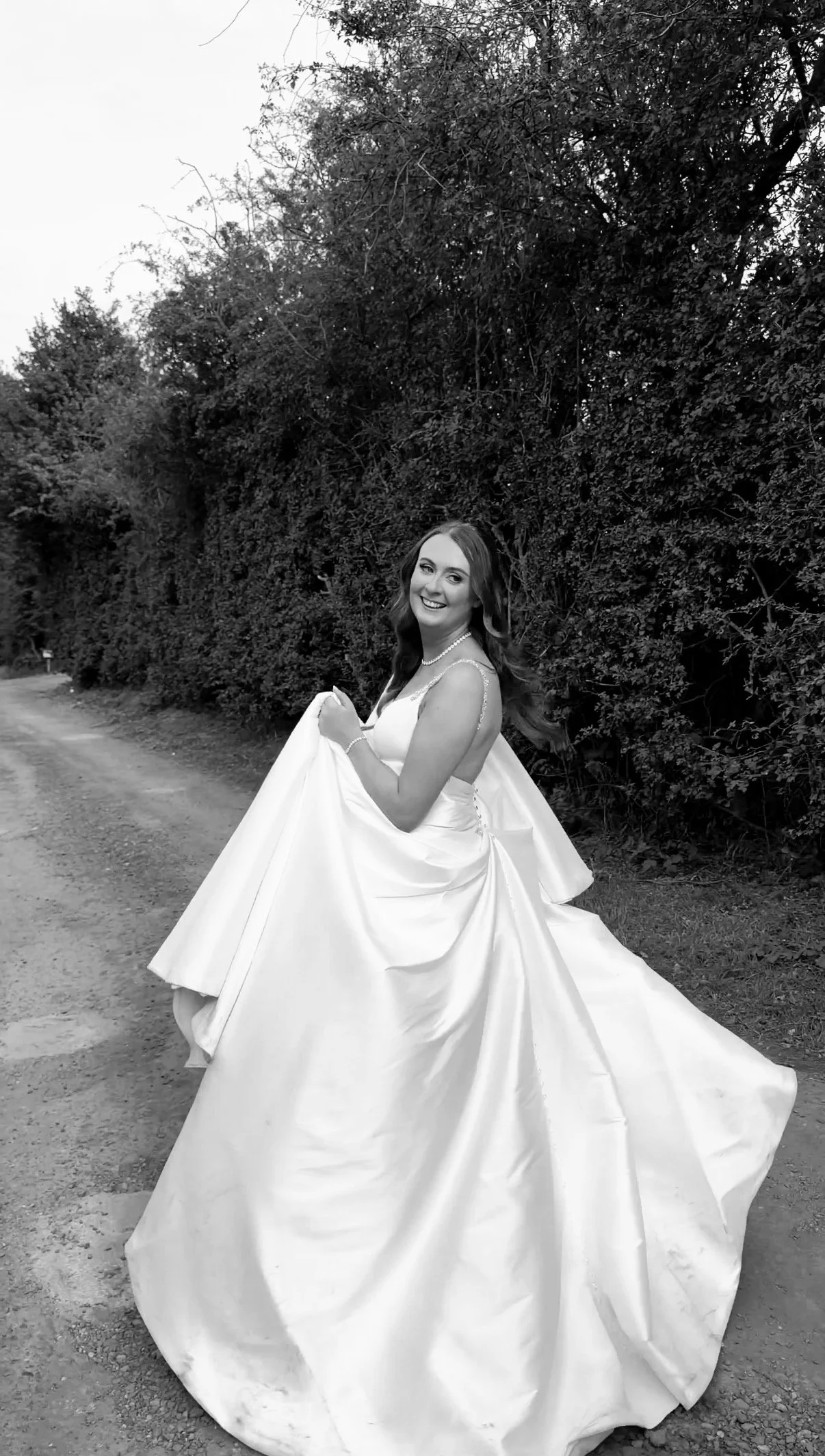 A woman in a wedding dress smiling outdoors next to a hedge.