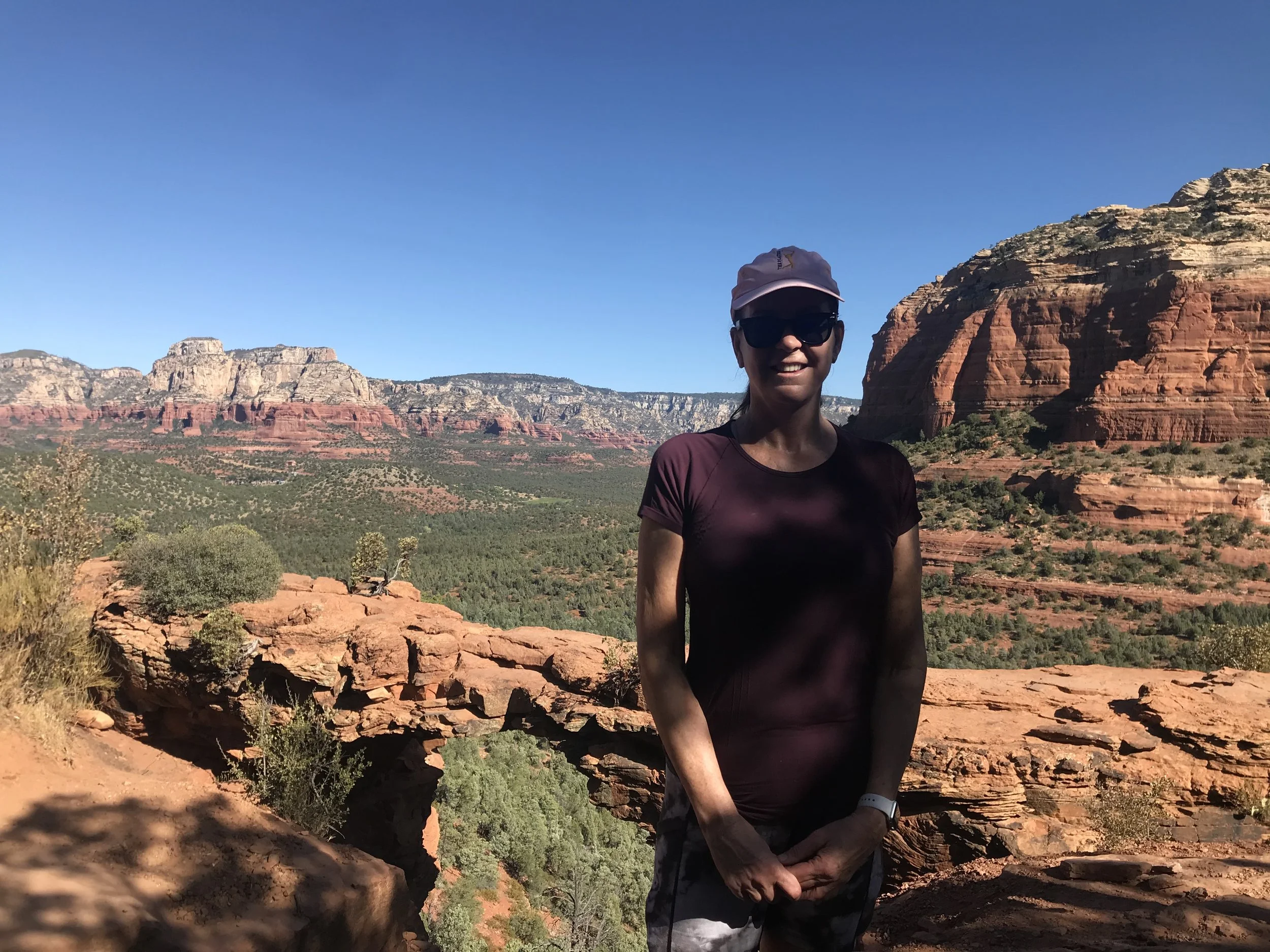 A person wearing a white hat, black sunglasses, a maroon shirt, and a watch, standing outdoors in front of layered red rock formations and a clear blue sky, possibly in a desert or national park.