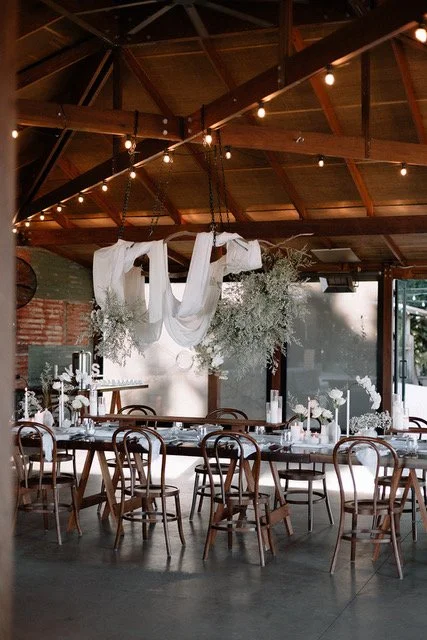Interior of a rustic event space with a long table, string lights, and a hanging fabric decoration with white flowers.