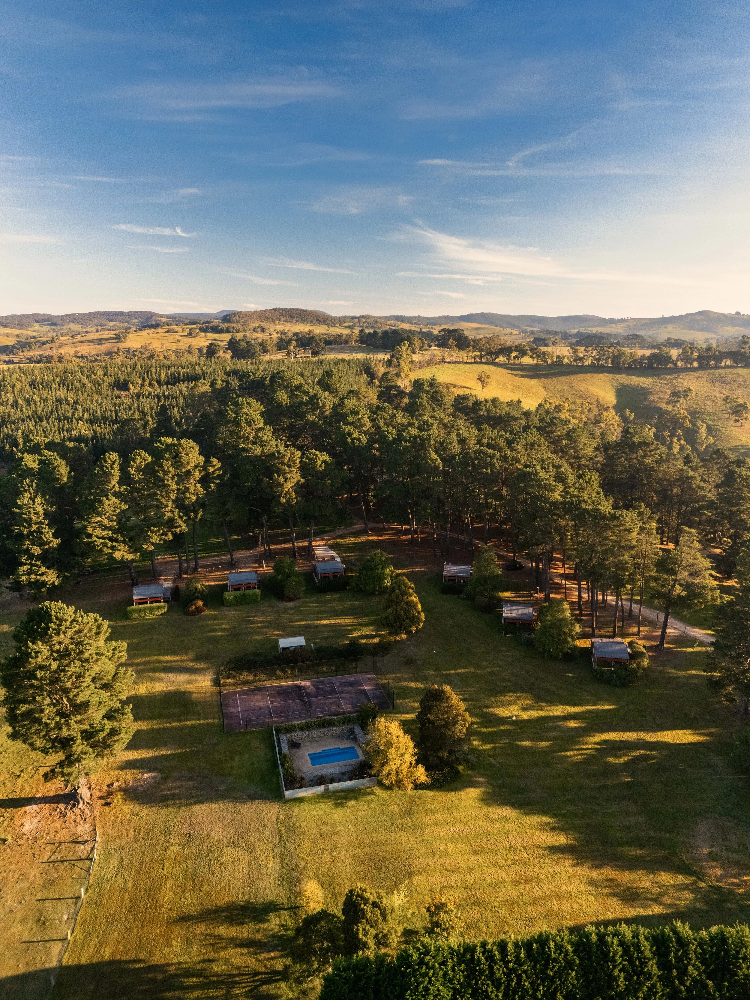 A scenic landscape with rolling hills, a dense forest, a tennis court, and a swimming pool, under a clear sky with wispy clouds.