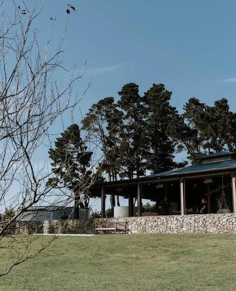 A modern outdoor patio with a stone wall, a covered roof, and an open-air seating area, surrounded by tall trees and a clear blue sky.