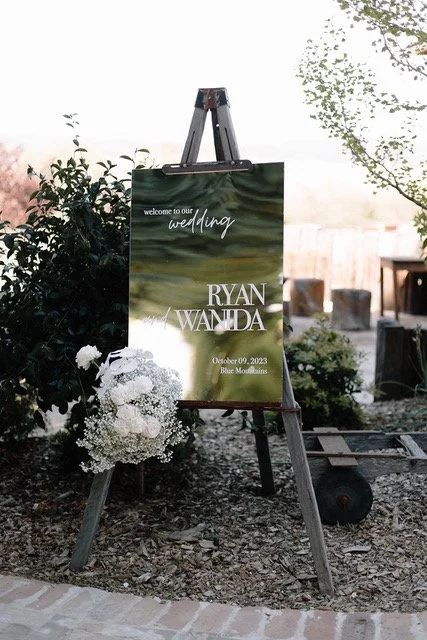 Wedding sign on an easel outdoors announcing Ryan and Wanda's wedding on October 9, 2023, at Blue Mountains with floral decorations nearby.