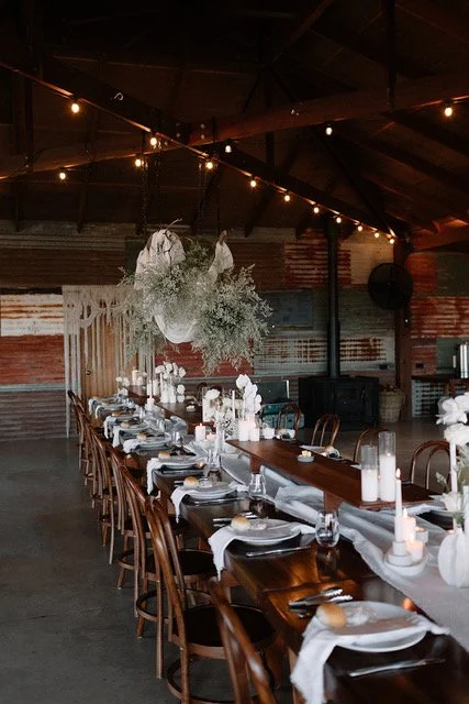 Long dining table set with white linens, candles, and floral centerpieces in a rustic industrial setting with string lights hanging overhead.