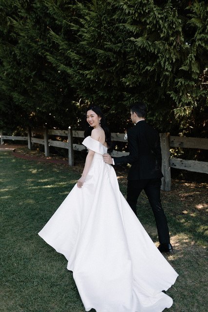 A woman in a white wedding gown and a man in a black tuxedo are standing outdoors on grass in front of trees and a wooden fence. The woman is smiling at the camera while holding the man's hand.