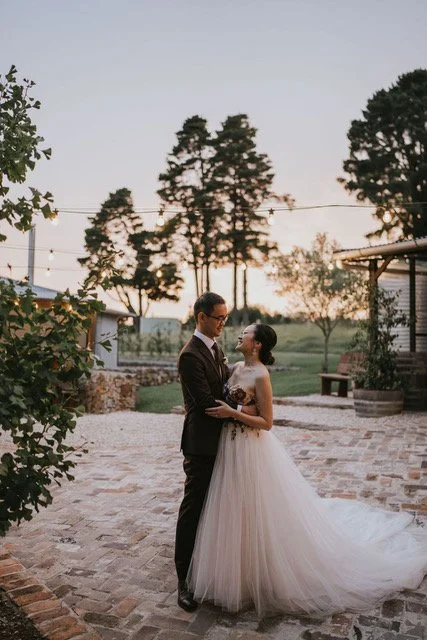 A bride and groom standing close together outside during sunset, with trees and string lights in the background.
