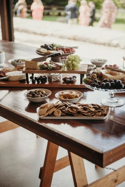 A table with various plates of cheese, crackers, grapes, olives, and other snacks, set in a room with a view of a garden or outdoor area with women dressed in pink in the background.