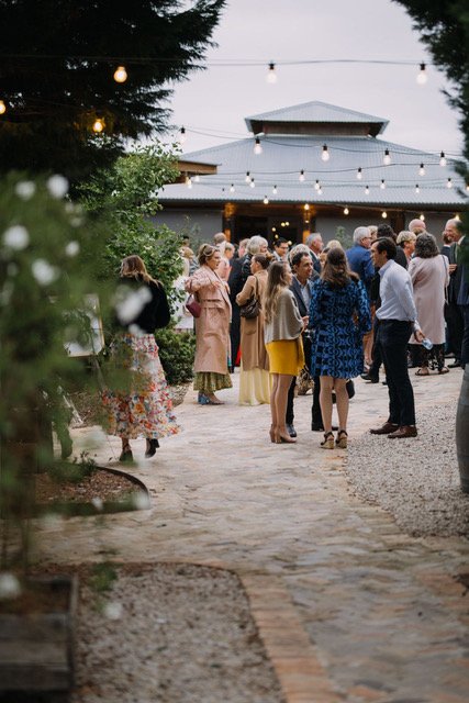 A gathering of people socializing outdoors during the evening, with string lights overhead and a pavilion in the background.