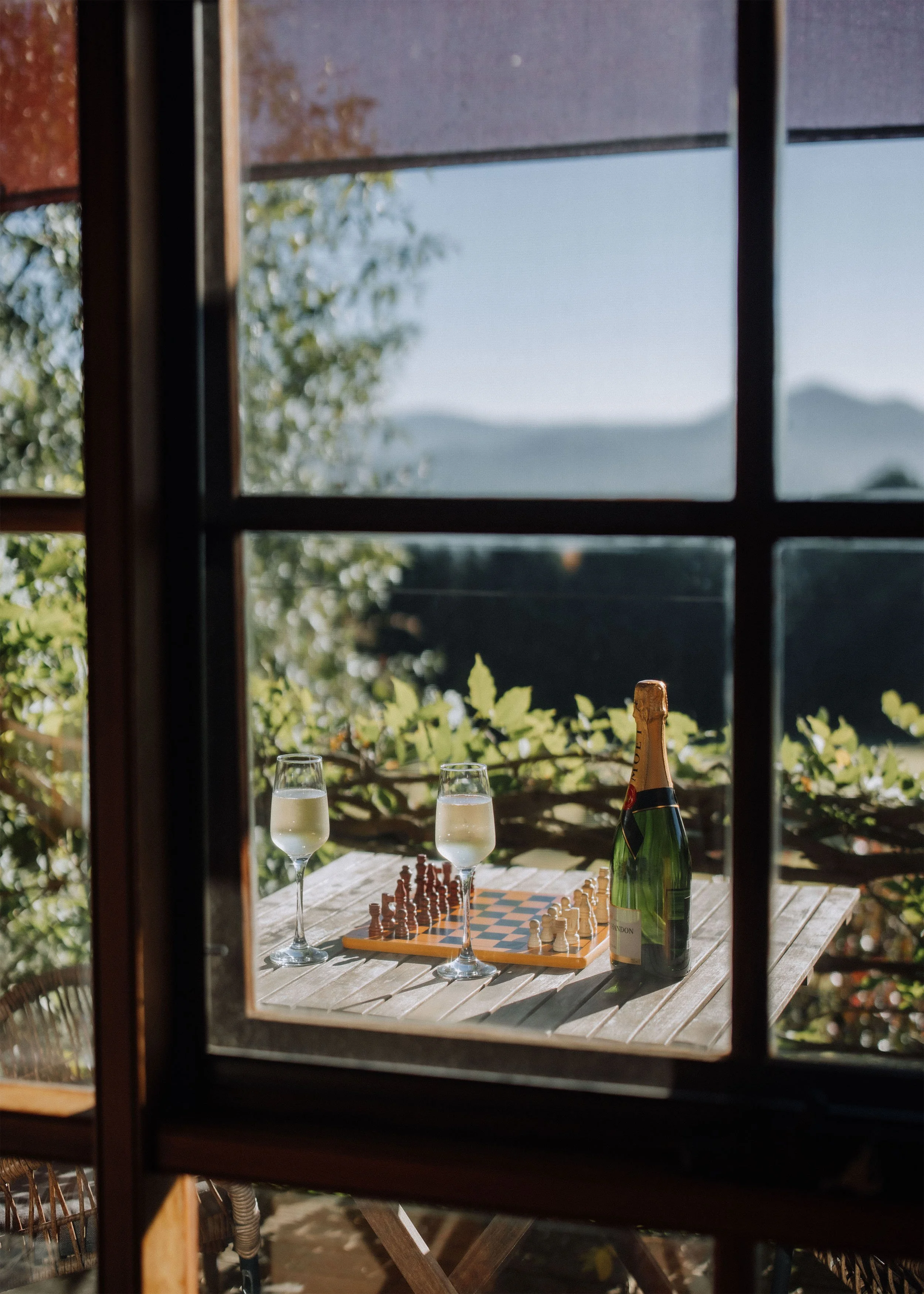 View through a window showing a wooden table with two glasses of champagne, a chessboard, and a bottle of champagne, with a mountainous landscape in the background.