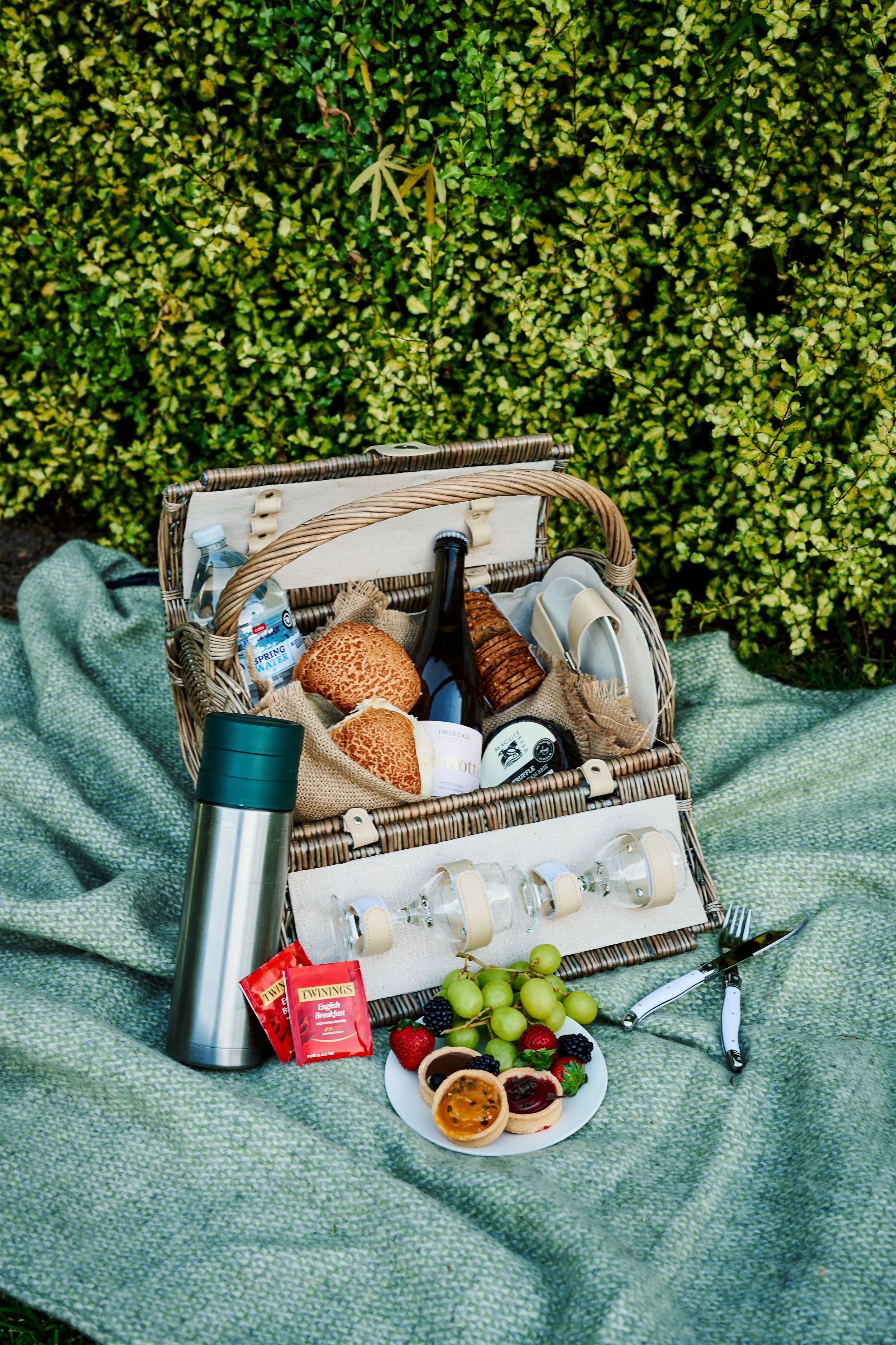 A picnic setup with a wicker basket containing bread, a bottle of wine, and various food items, placed on a green blanket in front of a hedge.