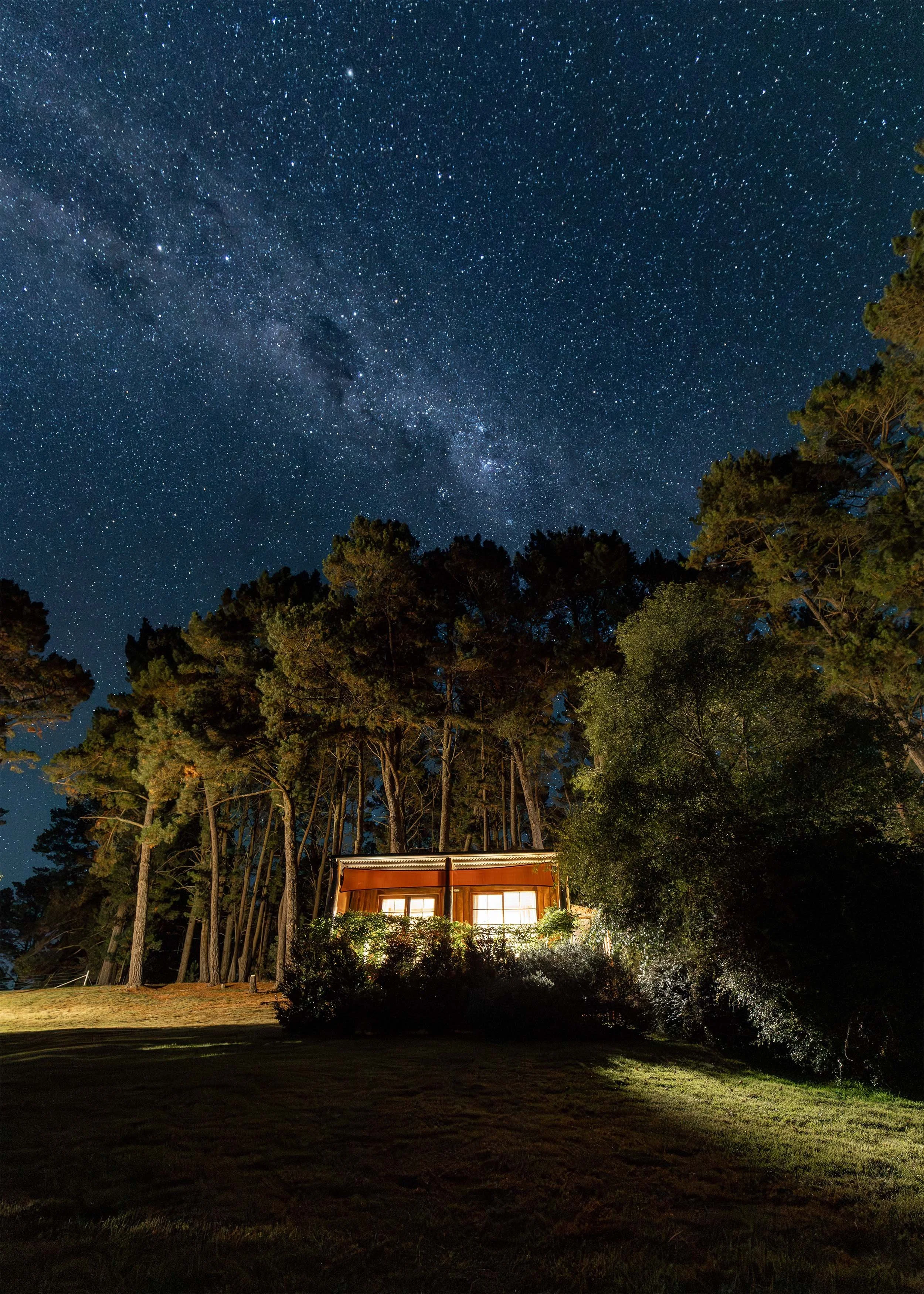 Nighttime scene showing a star-filled sky with the Milky Way galaxy visible. In the foreground, there is a small house with illuminated windows surrounded by trees and bushes.