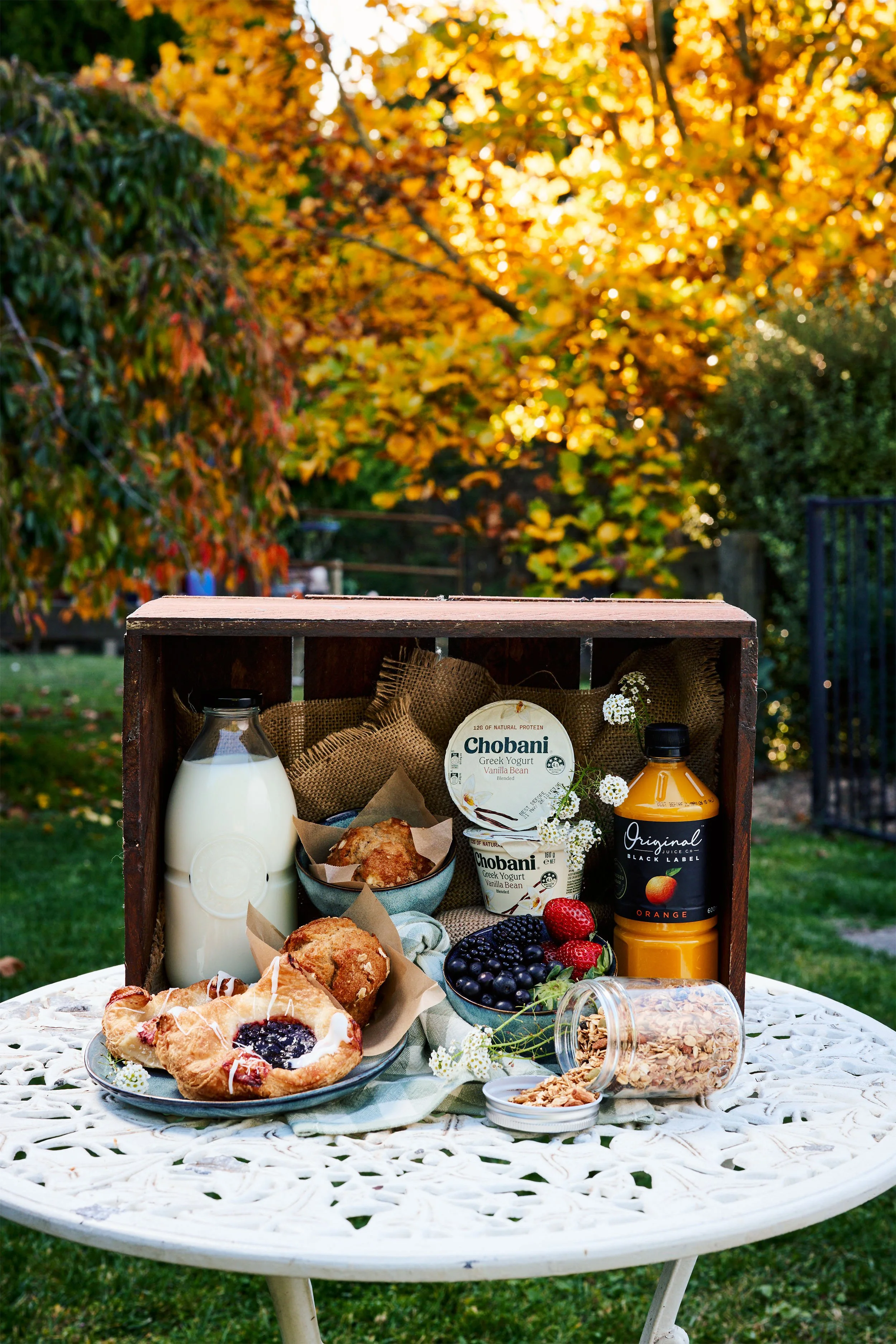 A picnic table set outdoors with breakfast foods, including pastries, berries, yogurt, a bottle of milk, a jar of oats, and a bottle of orange juice, with a background of trees with yellow and green leaves.