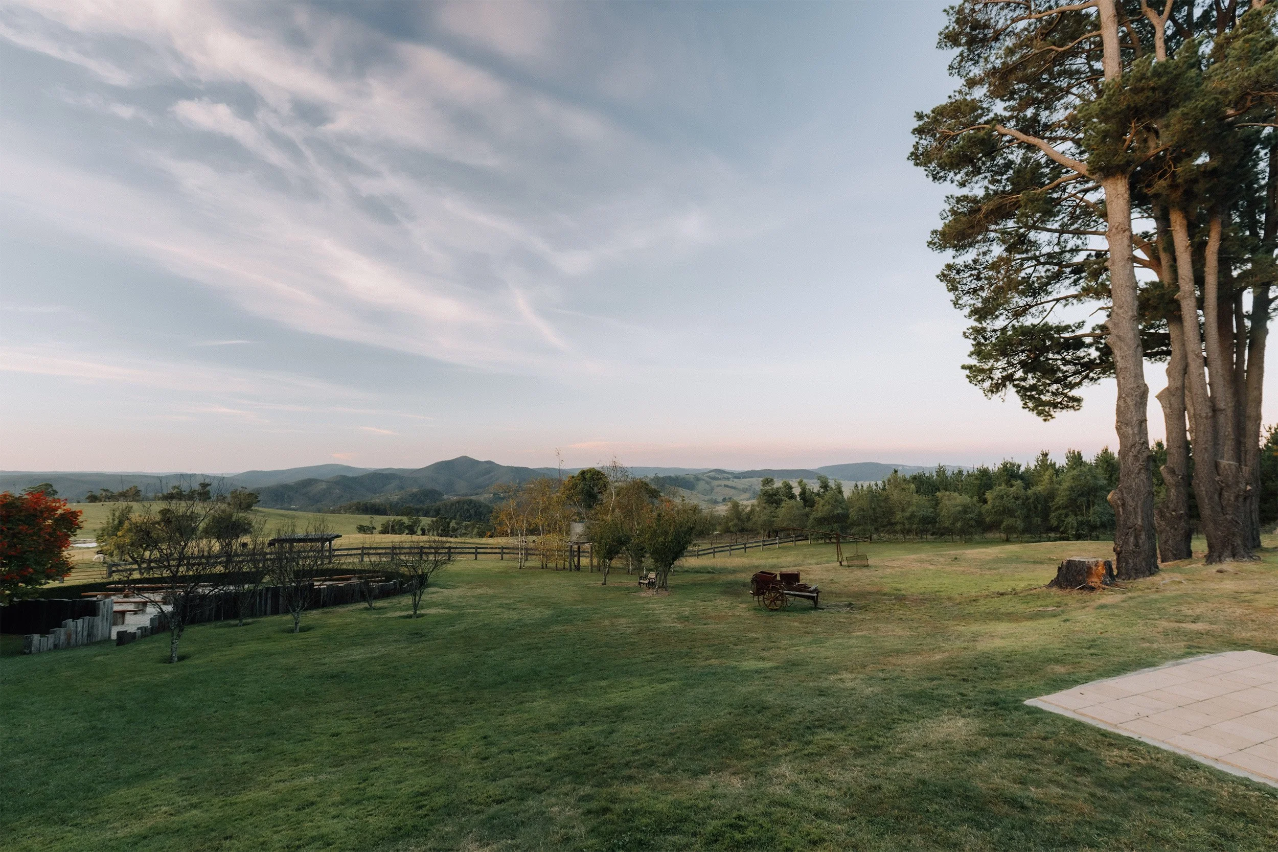 A peaceful rural landscape with green grass, large trees, a wooden fence, and distant mountains under a cloudy sky during early evening.