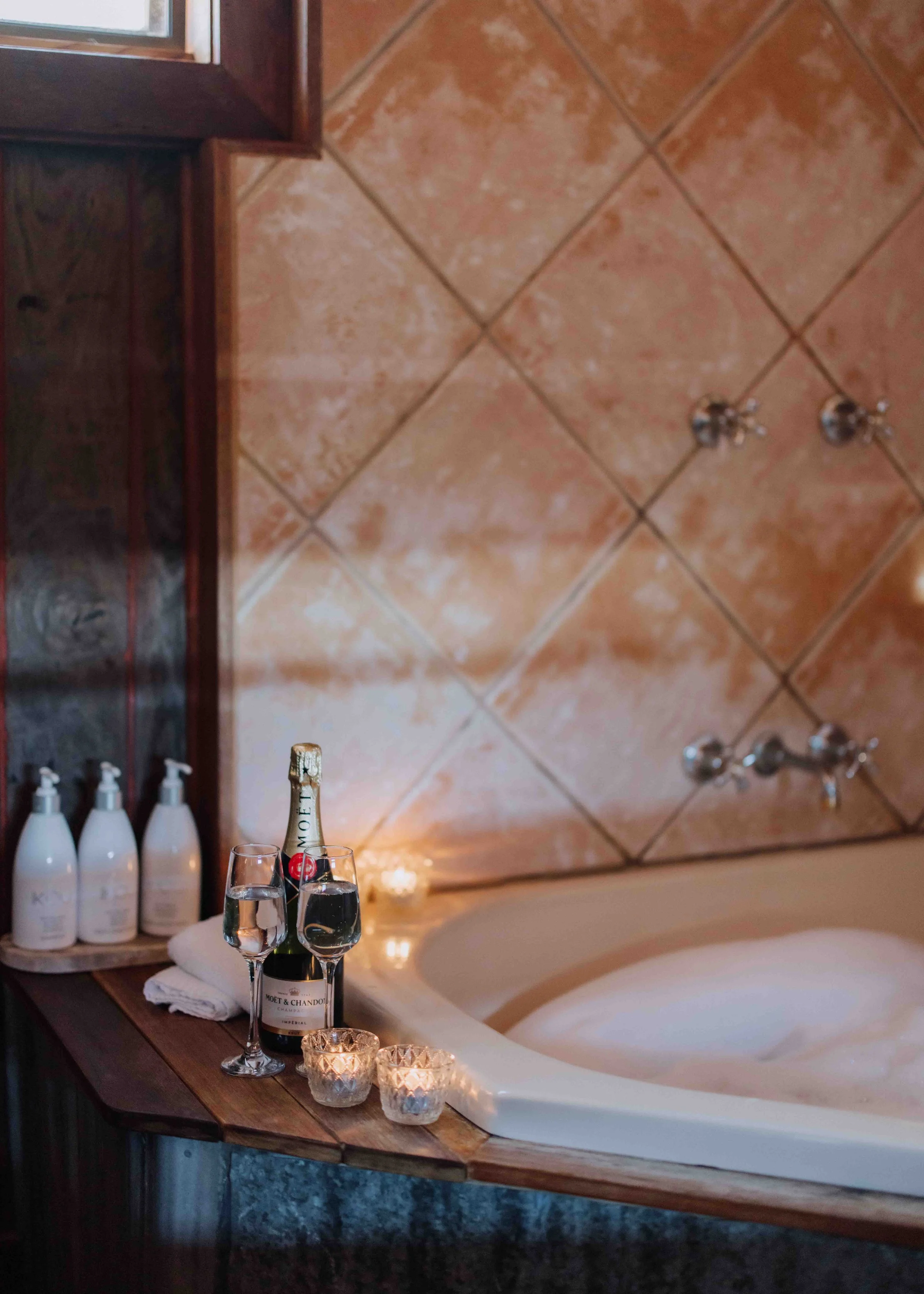 A bathroom scene featuring a bathtub with candles and drinks on a wooden ledge, bottles of toiletries, a bottle of Moët & Chandon champagne, two glasses of water, and a partially visible window with tile walls in the background.
