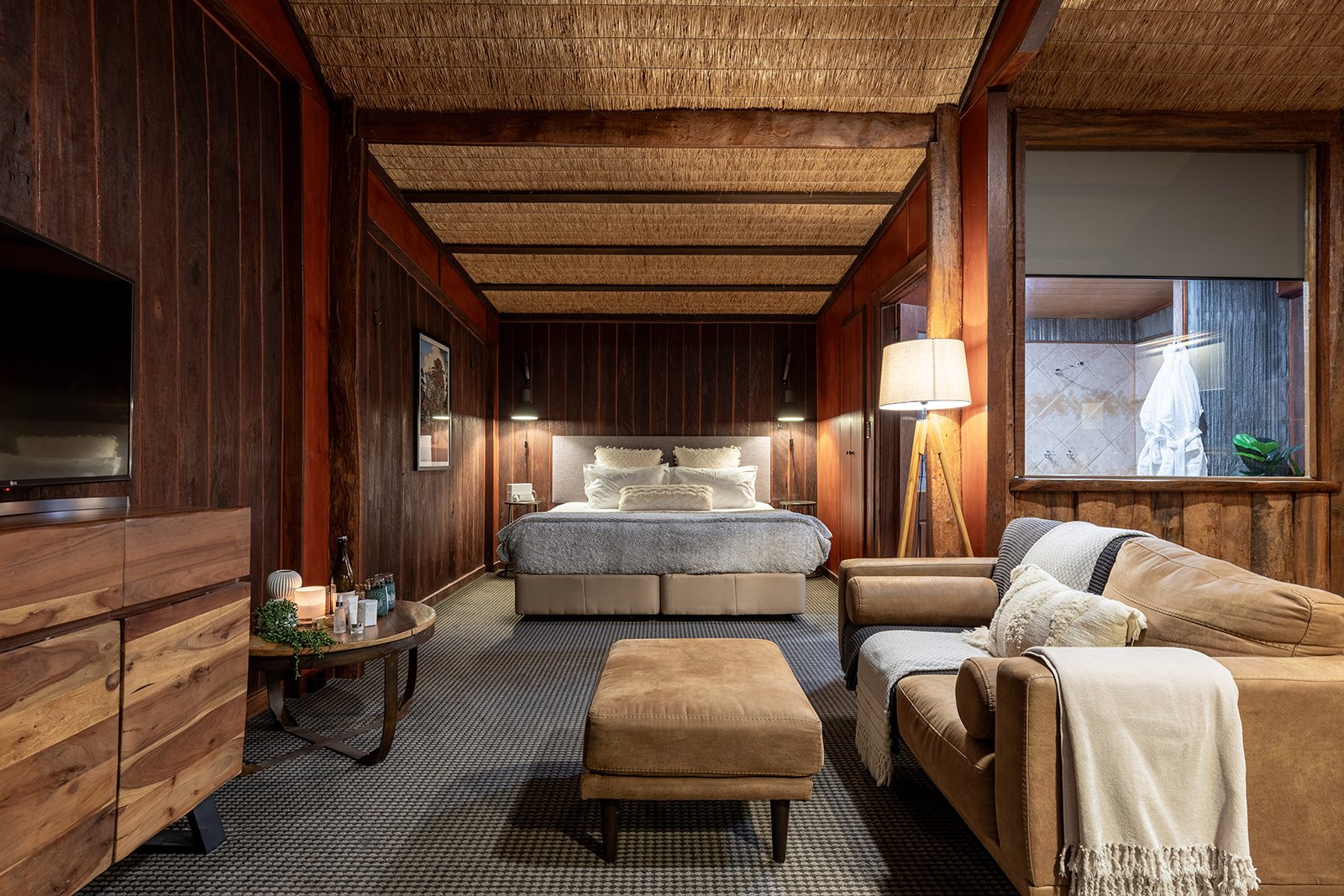 A cozy hotel room with wood-paneled walls, a large bed with grey and white bedding, a beige armchair, a matching ottoman, a wooden dresser, and a window with a view of a balcony with a bathrobe hanging outside.
