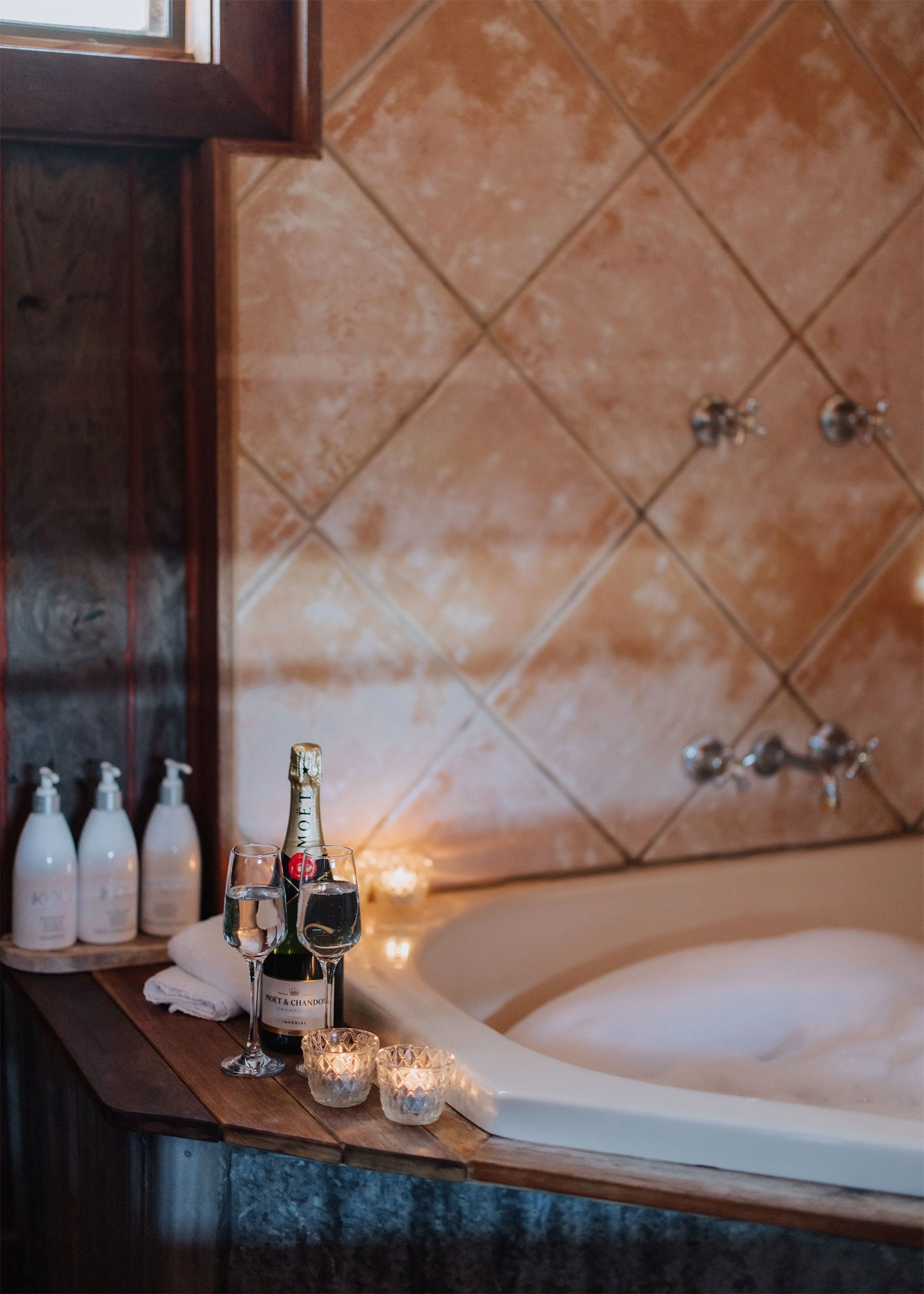 A bathroom with a bathtub, lit candles, two glasses of champagne, and a bottle of Moët & Chandon champagne. There are bottles of toiletries on a wooden shelf, and a tiled wall with two faucets.