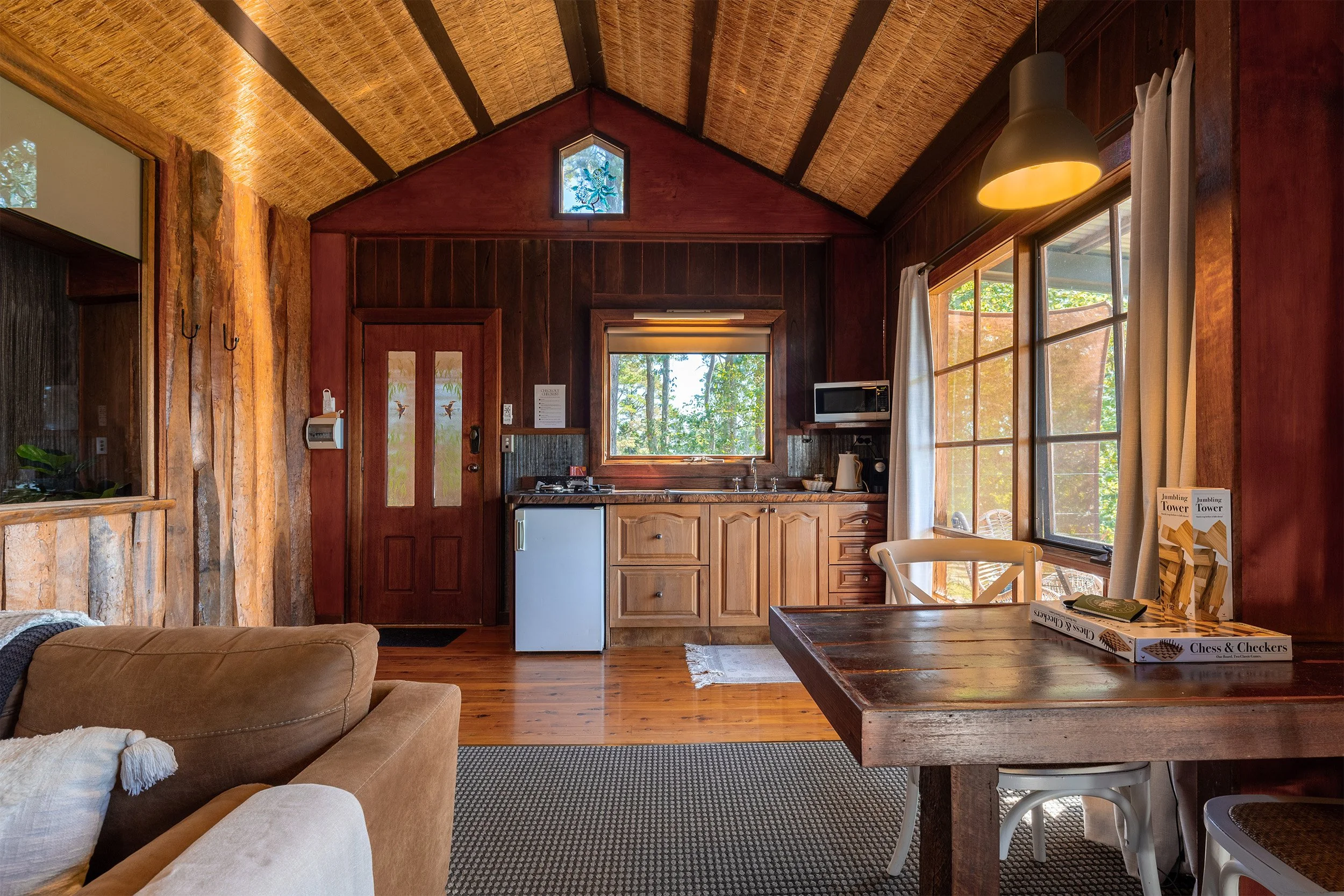 Interior view of a cozy wooden cabin kitchen and living space with a sloped wooden ceiling, large windows with curtains, a small refrigerator, microwave, and a wooden dining table with books on top. Natural light filters through greenery outside.