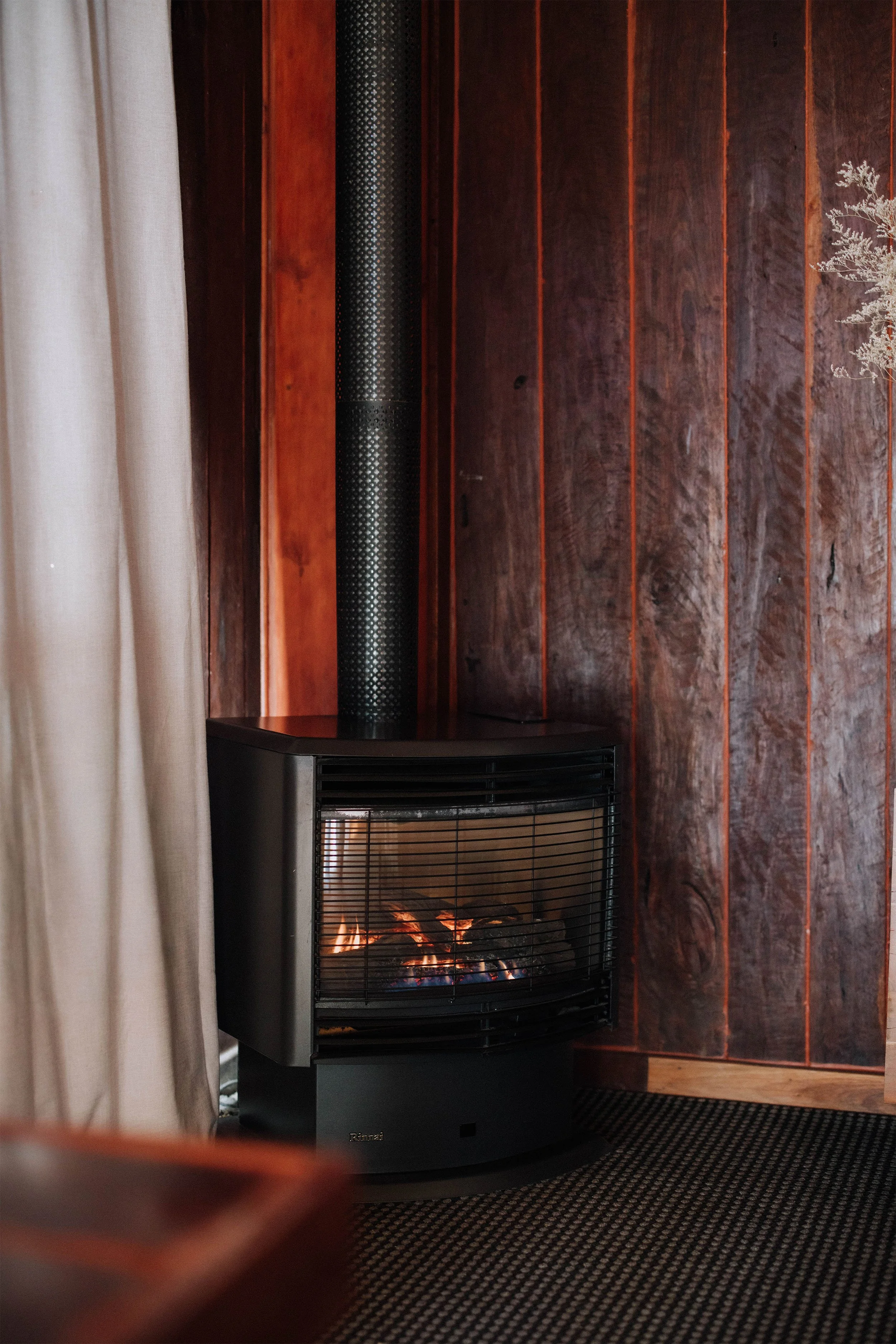 A fireplace with a fire burning inside, placed against a wooden wall with vertical panels. To the left, part of a light-colored curtain is visible, and the edge of a black object is seen in the foreground.