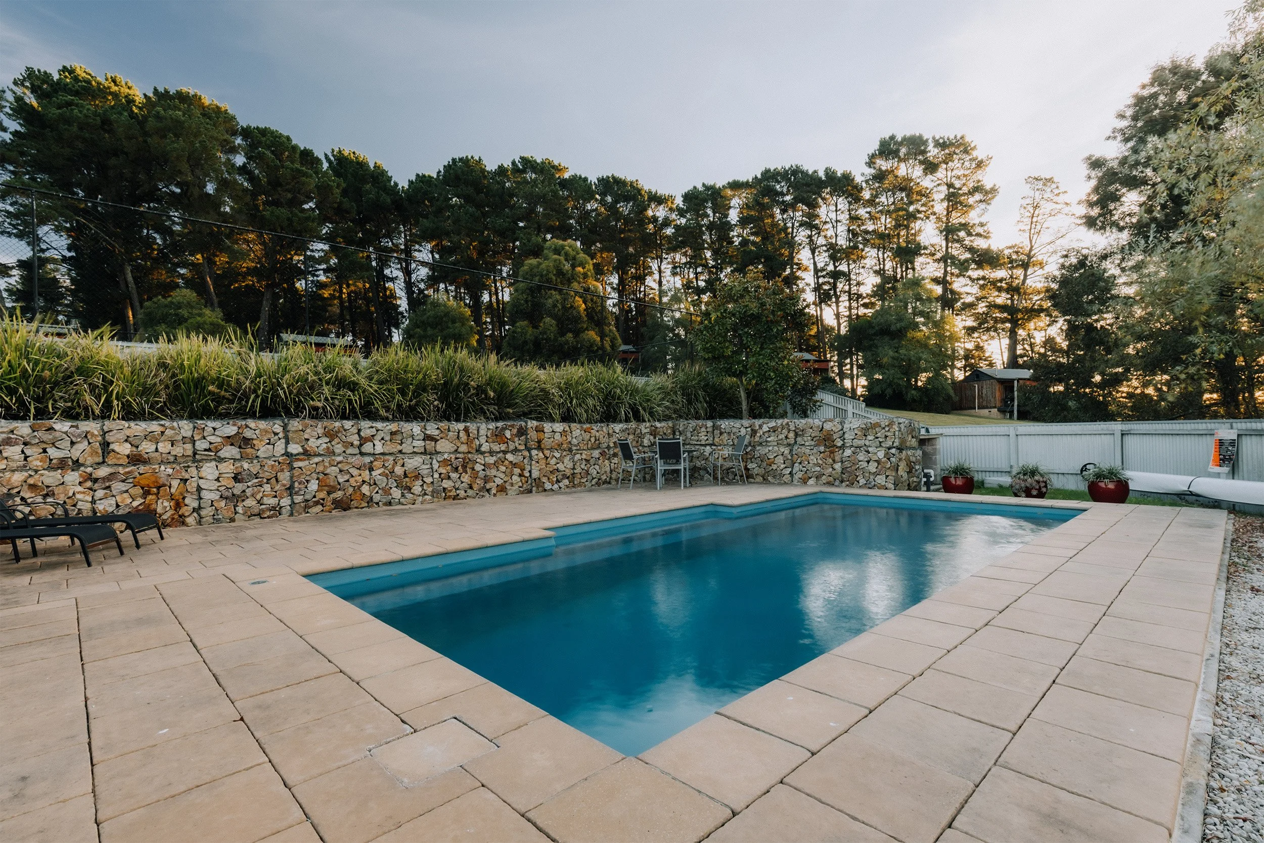 A backyard with a swimming pool, surrounded by stone patio and chairs, and a tall trees backdrop during sunset.