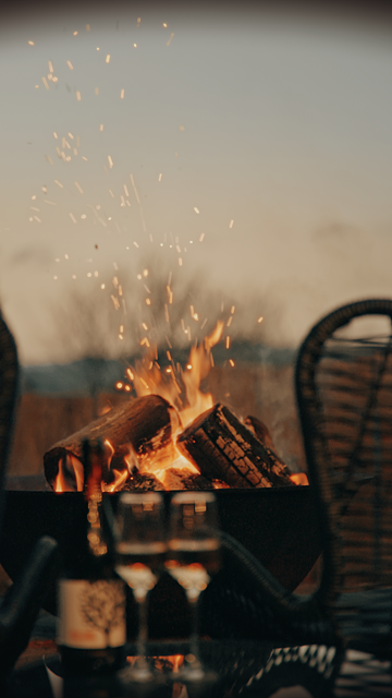 Campfire with burning logs and sparks, surrounded by outdoor chairs and a table with drinks.