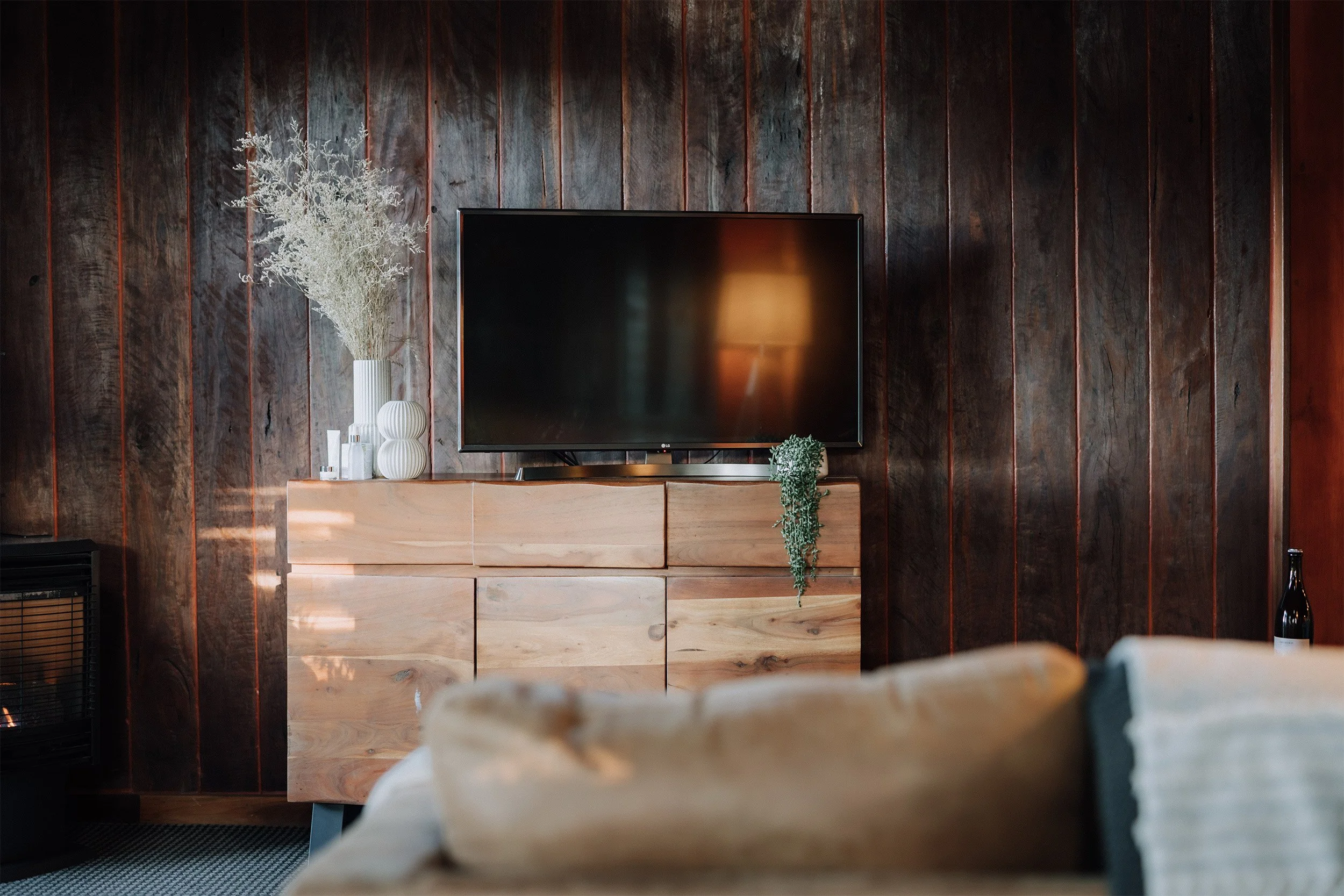 A flat-screen TV on a wooden cabinet against a dark wood-paneled wall, with decorative vases, a plant, and a lamp reflected on the screen.