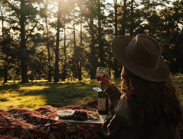 Person wearing a wide-brimmed hat sitting on a blanket in a forested area during sunset, holding a glass of wine with a bottle nearby, and a plate with food.