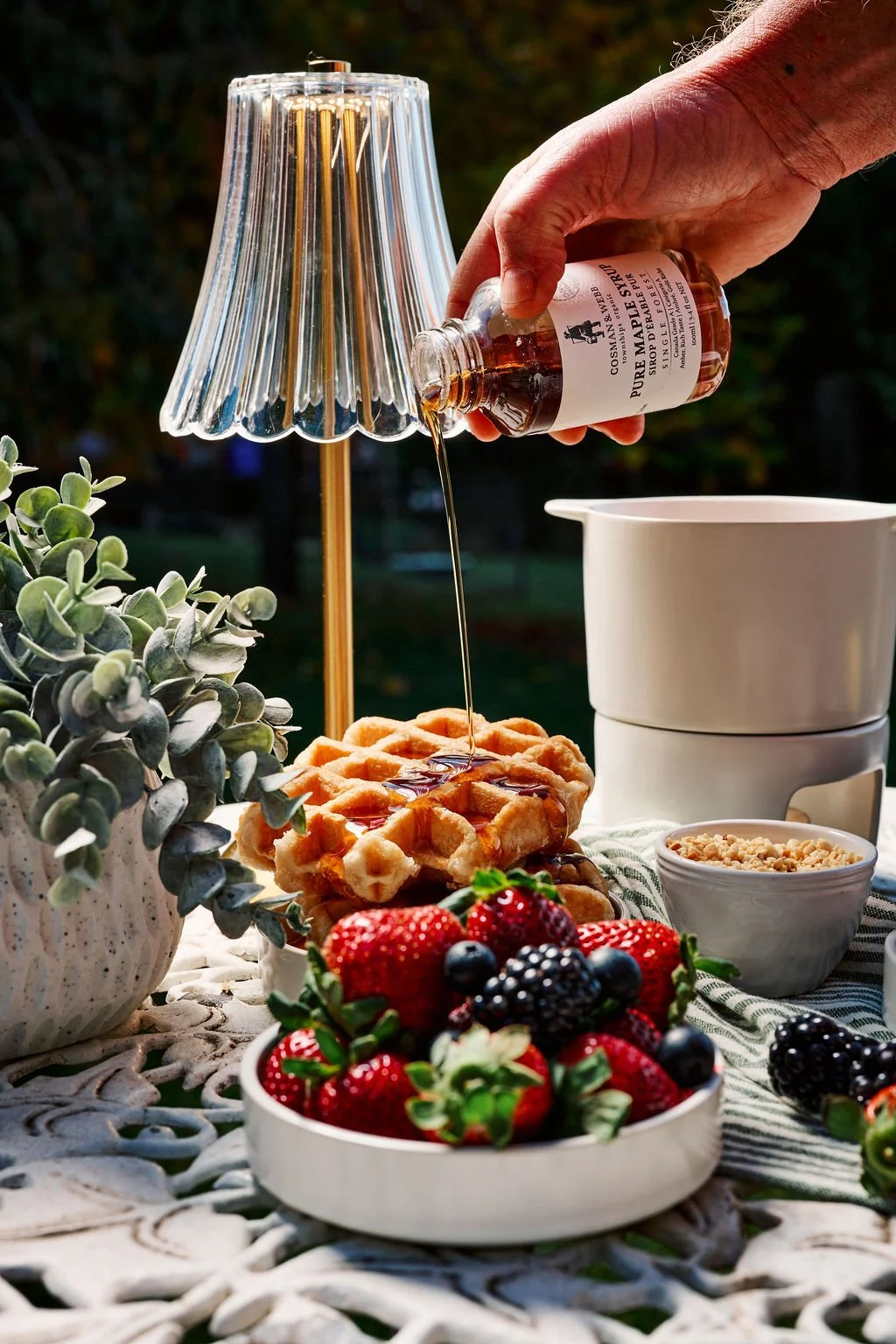A person pouring syrup from a glass jar onto a golden waffle topped with berries and syrup, with a bowl of mixed berries, a potted plant, and white dishes on a lace tablecloth outdoors in natural light.