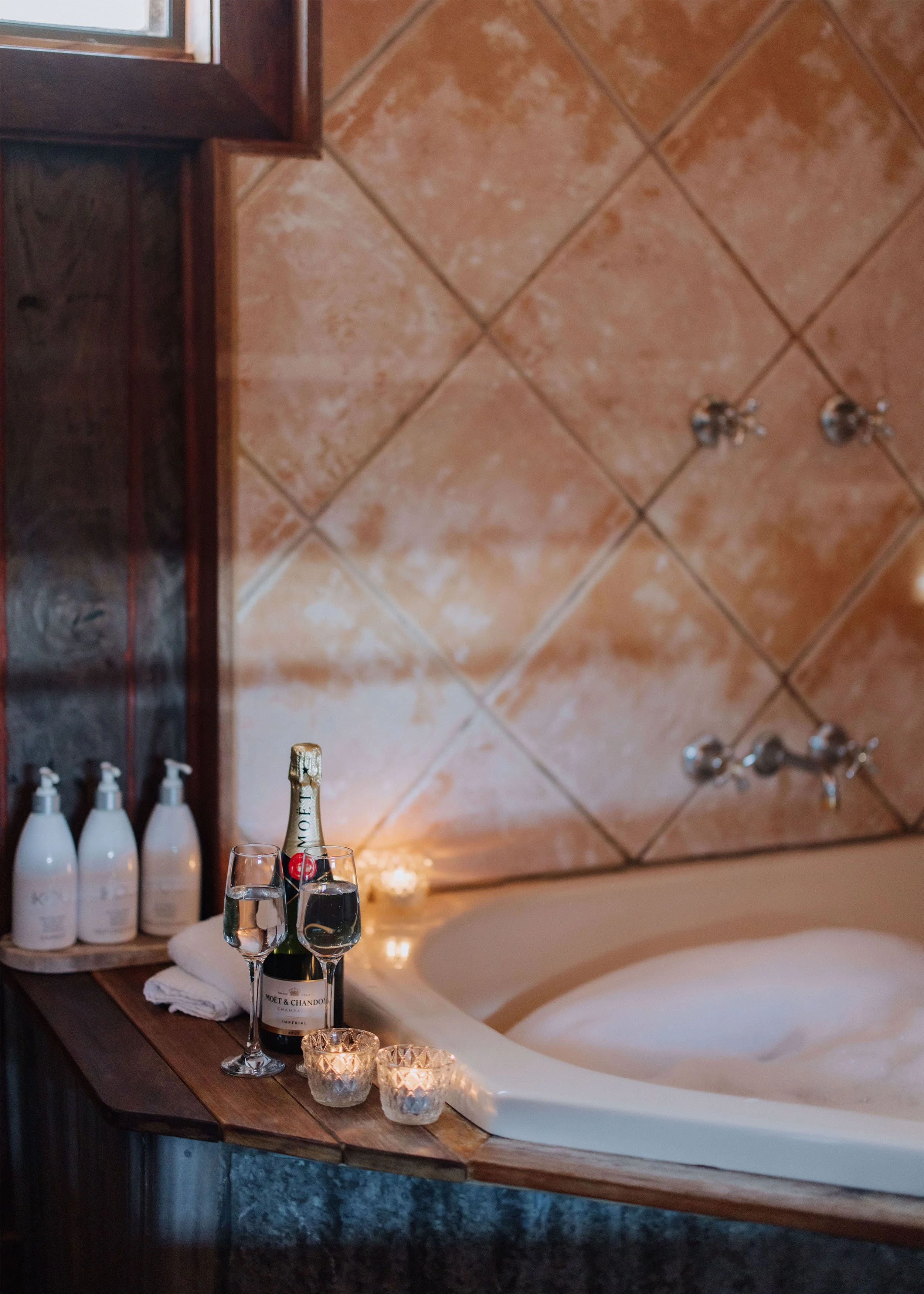 A bathroom scene with a bathtub, a wooden shelf with four bottles of soap or lotion, two glasses of water, a bottle of Moët & Chandon champagne, and three lit candles.