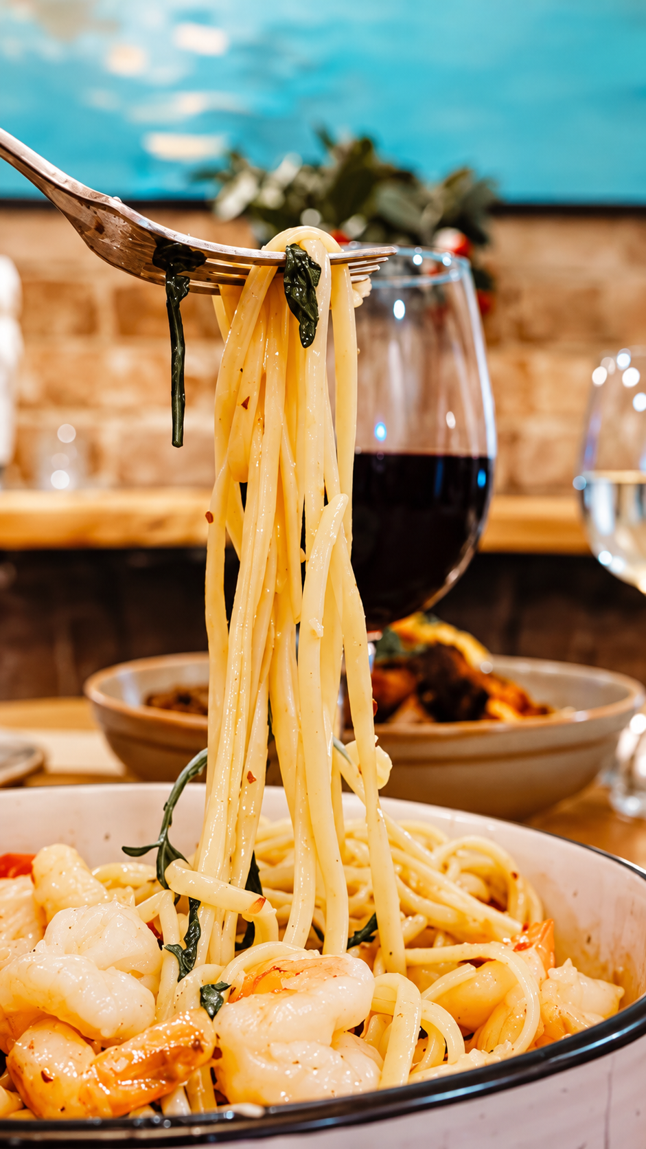 Spaghetti with shrimp and green herbs, a glass of red wine, and a bowl of food on a table in a cozy restaurant setting.